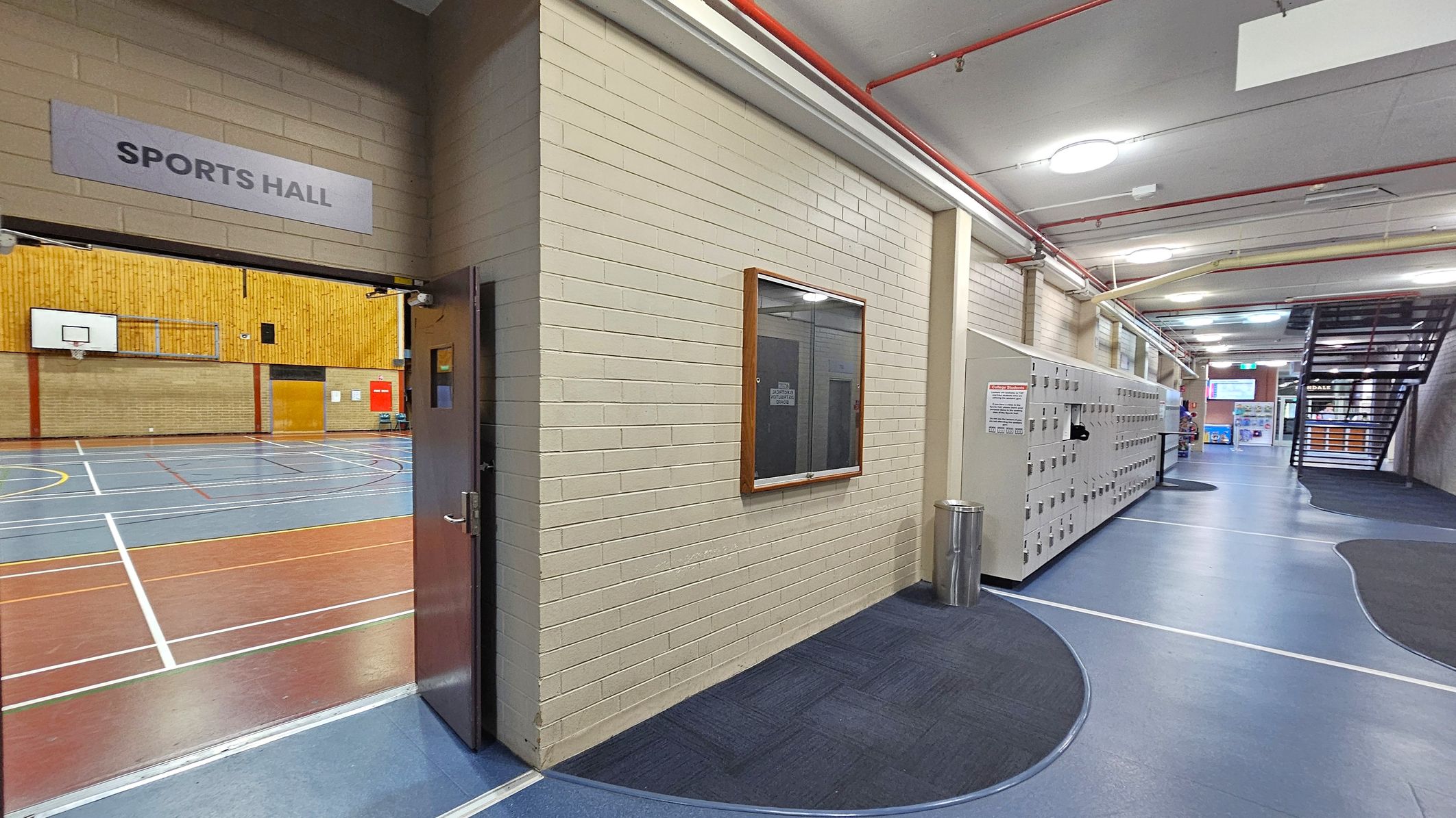 A corridor with a sports hall entrance on the left, marked by a sign above the door. Inside, a basketball hoop and court lines are visible. The right side features a row of lockers along a blue and grey floor. A notice board is mounted on the wall near a metal bin. The ceiling has exposed pipes and circular lights. In the background, a staircase leads upwards, and there are posters and signs near the exit.