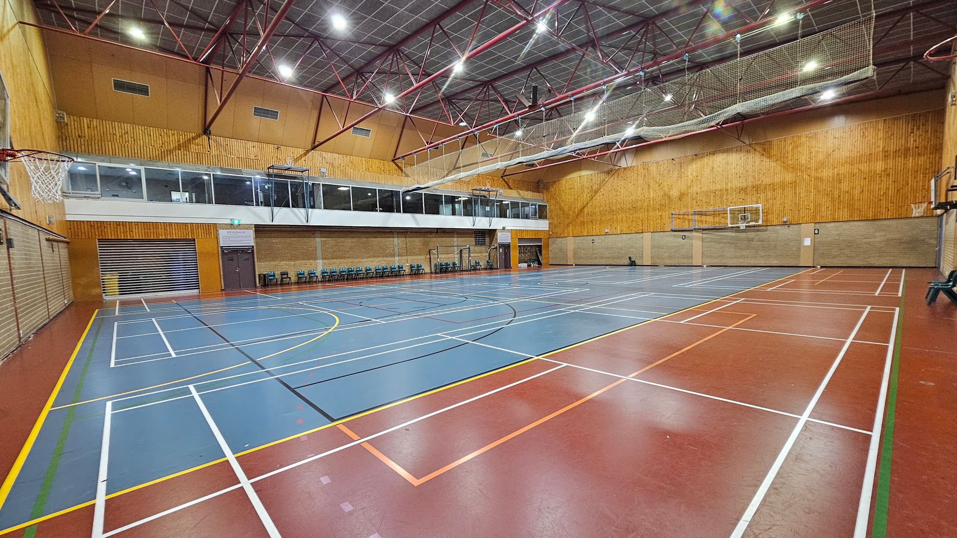 A spacious indoor sports hall with a blue and red floor marked for various sports. On the left, a basketball hoop is visible. The right side features another hoop and a brick wall. The back wall is lined with a row of blue chairs and a viewing gallery above. The ceiling is high with red metal beams and bright lights. The walls are a mix of wood panelling and brick.