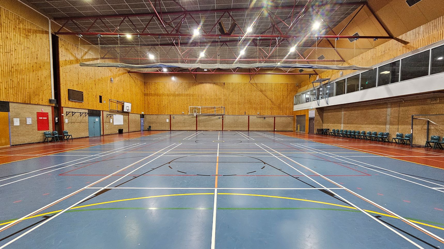 A spacious indoor sports hall with a blue floor marked with various sports lines in white, yellow, and orange. The walls are clad in light wood panelling. On the left, there are chairs and a red fire hose reel. The right side features a row of chairs against a brick wall and a large windowed viewing area above. The ceiling is high with exposed beams and lighting fixtures. A basketball hoop is visible at the far end.