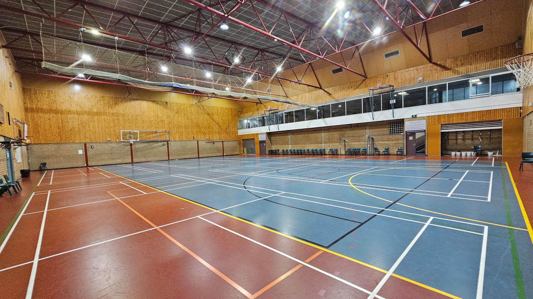 A spacious indoor sports hall with a high ceiling supported by red metal beams. The floor is marked with various sports lines, predominantly blue and red. On the right, there are rows of chairs against the wall, and a basketball hoop is visible. The left side features more chairs and another basketball hoop. The walls are a mix of wood panelling and brick. Large windows and a viewing area are situated above the chairs on the right side. The ceiling has a netting structure.