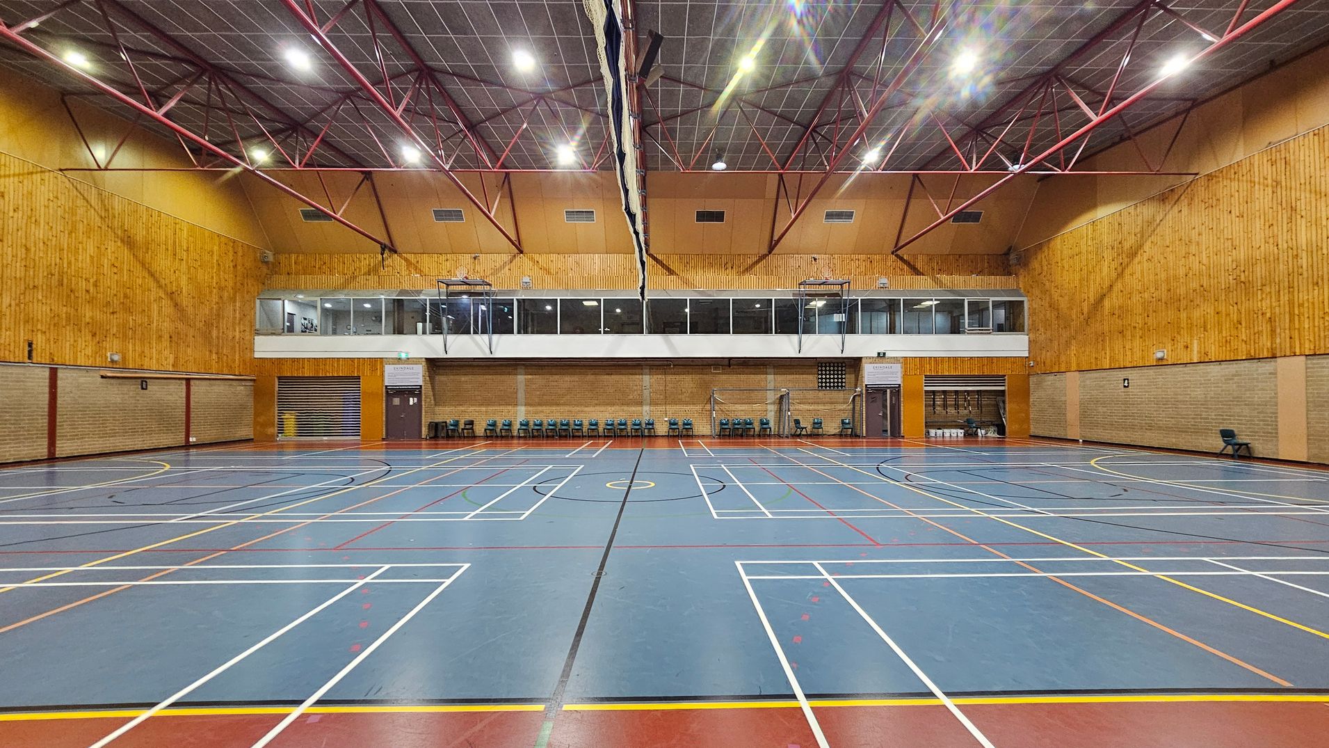 A spacious indoor sports hall with a blue floor marked for various sports. The walls are clad in wood panelling, and there are rows of chairs lined up against the far wall. Above, a mezzanine level with large windows overlooks the hall. The ceiling is high with exposed red metal beams and bright lighting. A net hangs down the centre of the hall.