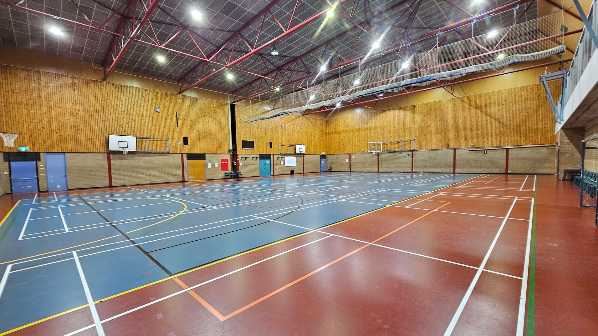 A spacious indoor sports hall with a high ceiling supported by red metal beams. The floor is marked with various sports lines in white, yellow, and orange on a blue and red surface. On the left, there are basketball hoops and blue doors. The walls are a mix of brick and wood panelling. On the right, a row of stacked chairs is visible. The ceiling features bright overhead lights and a netting system.