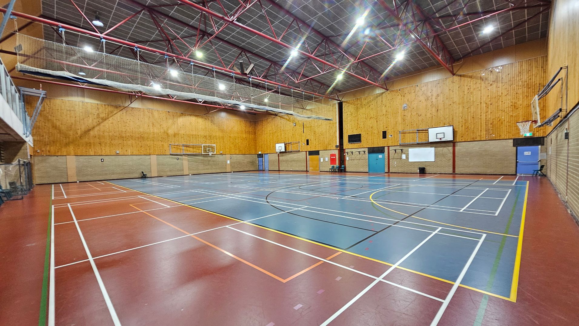A spacious indoor sports hall with a wooden floor marked for various sports. On the left, there is a red section with white lines. The centre features a blue area with additional markings. The walls are made of light brown brick and wood panelling. Several basketball hoops are mounted on the walls. The ceiling is high with red metal beams and bright lighting. A net is suspended above the court. Doors in blue and orange are visible on the right wall.
