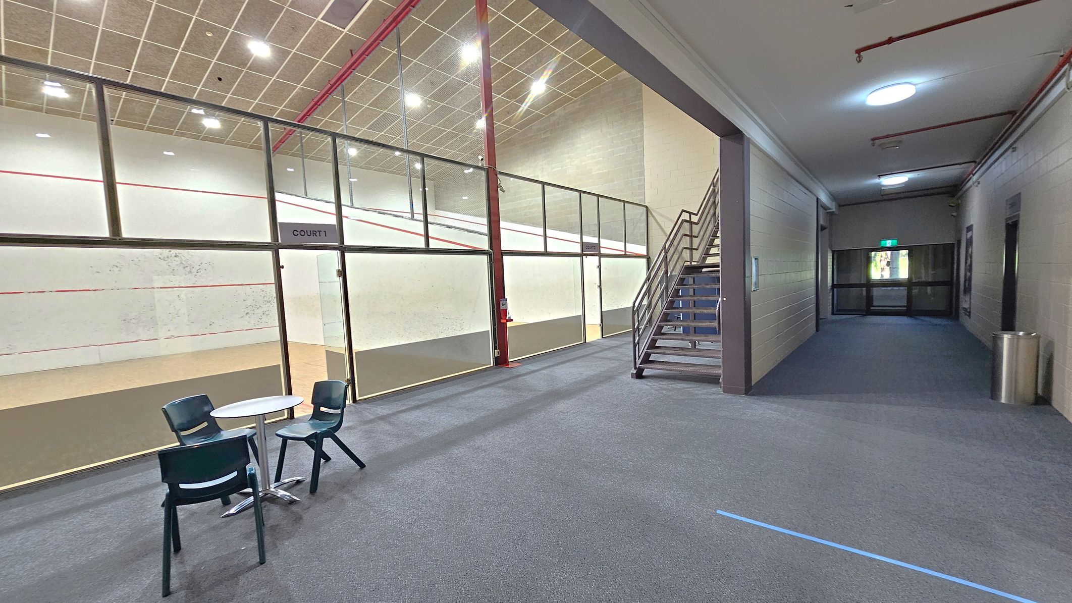 A squash court is visible on the left, enclosed by glass walls with a sign reading "Court 1". In front of the court, there is a small round table with two chairs. To the right, a staircase with metal railings leads upwards. The corridor extends to the right, with a blue carpeted floor and grey walls. The ceiling features a grid pattern with recessed lighting. A metal bin is positioned near the corridor's entrance.