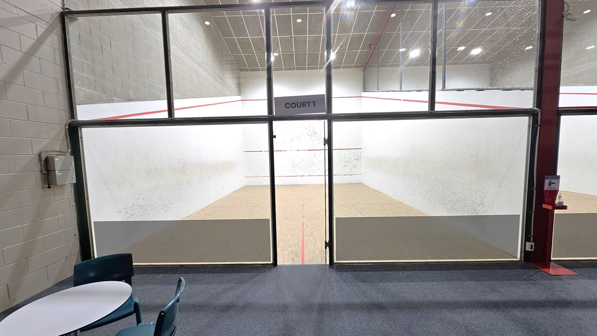 A squash court is visible through large glass doors labelled "Court 1". The court has white walls with red boundary lines and a wooden floor. In the foreground, there is a small round table with blue chairs on a carpeted floor. The ceiling is high with a grid pattern and bright lights. The left wall is made of grey bricks, and there is a hand sanitiser dispenser mounted on the right side near the entrance.