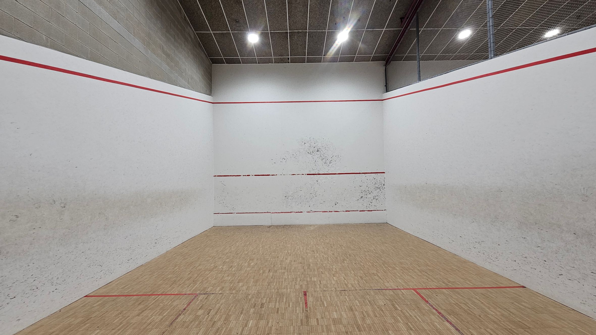 A squash court with white walls marked by red lines. The front wall has visible scuff marks from play. The floor is wooden with red boundary lines. The ceiling is grey with a grid pattern and several bright lights. The side walls are also white, showing signs of wear.