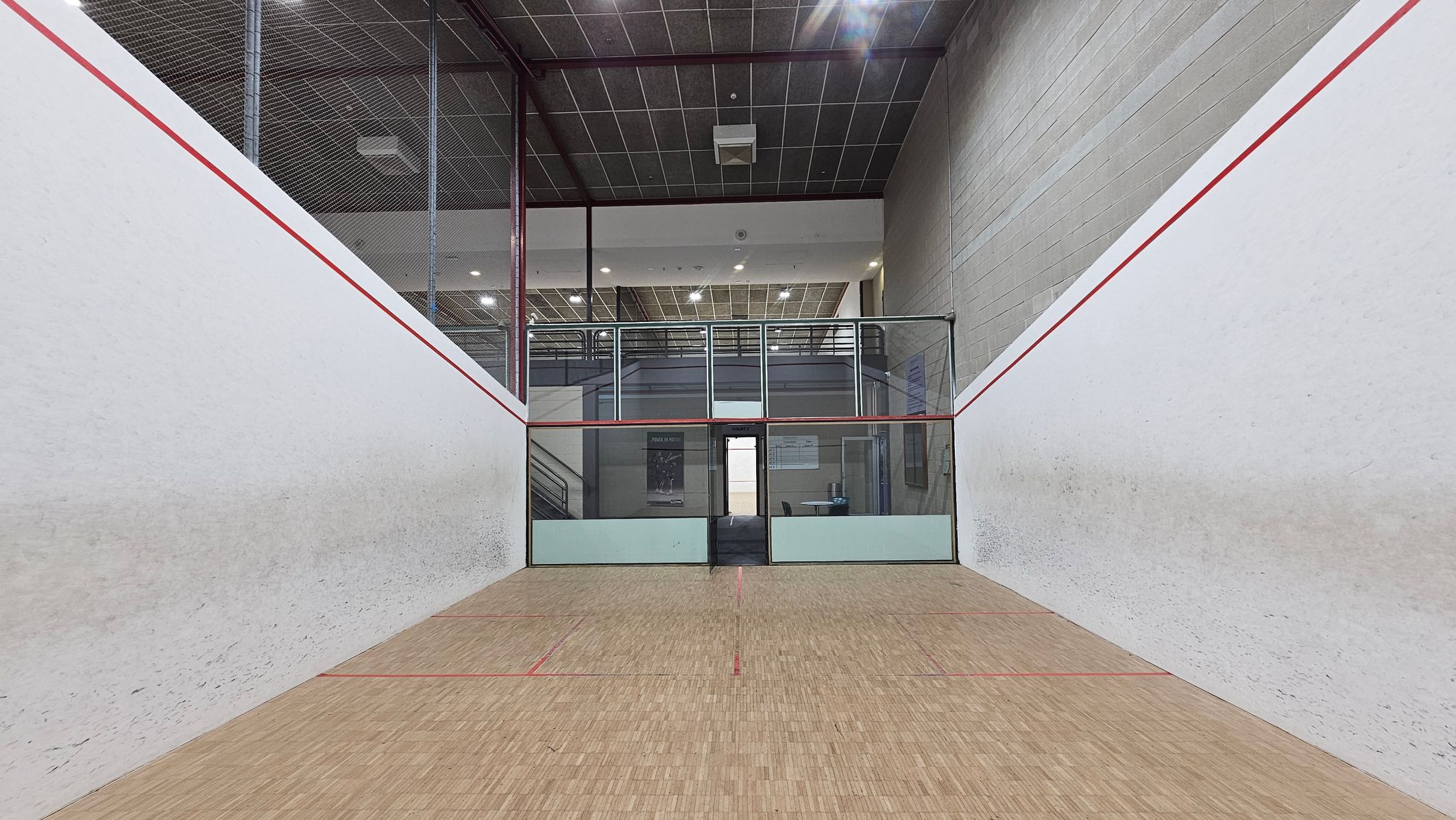 A squash court with white walls and red boundary lines. The floor is wooden with red markings. At the far end, there is a glass wall with a door leading to a viewing area. Above the glass wall, there is a balcony with railings and a ceiling with a grid pattern and lights. The walls are slightly scuffed from play.