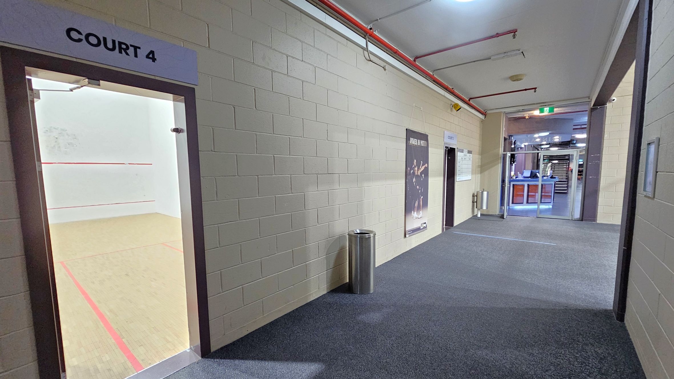 A corridor with a squash court entrance on the left labelled "Court 4". The court has a wooden floor with red boundary lines. The right side features a brick wall with a poster and a metal bin. Further down the corridor, there are more doors and a glass entrance leading to another area. The floor is carpeted, and the ceiling has exposed pipes and lighting fixtures.