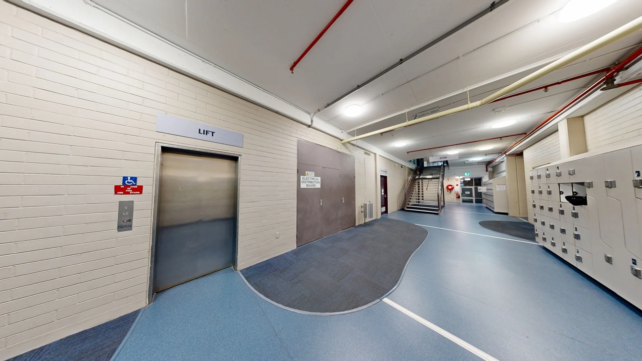 An elevator on the left and a staircase toward the right of the image along a wide corridor with blue vinyl flooring and white coloured brick walls.