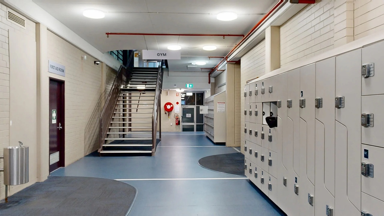 A staircase along a wide corridor with white brick walls and the right side wall having storage lockers on blue vinyl flooring.