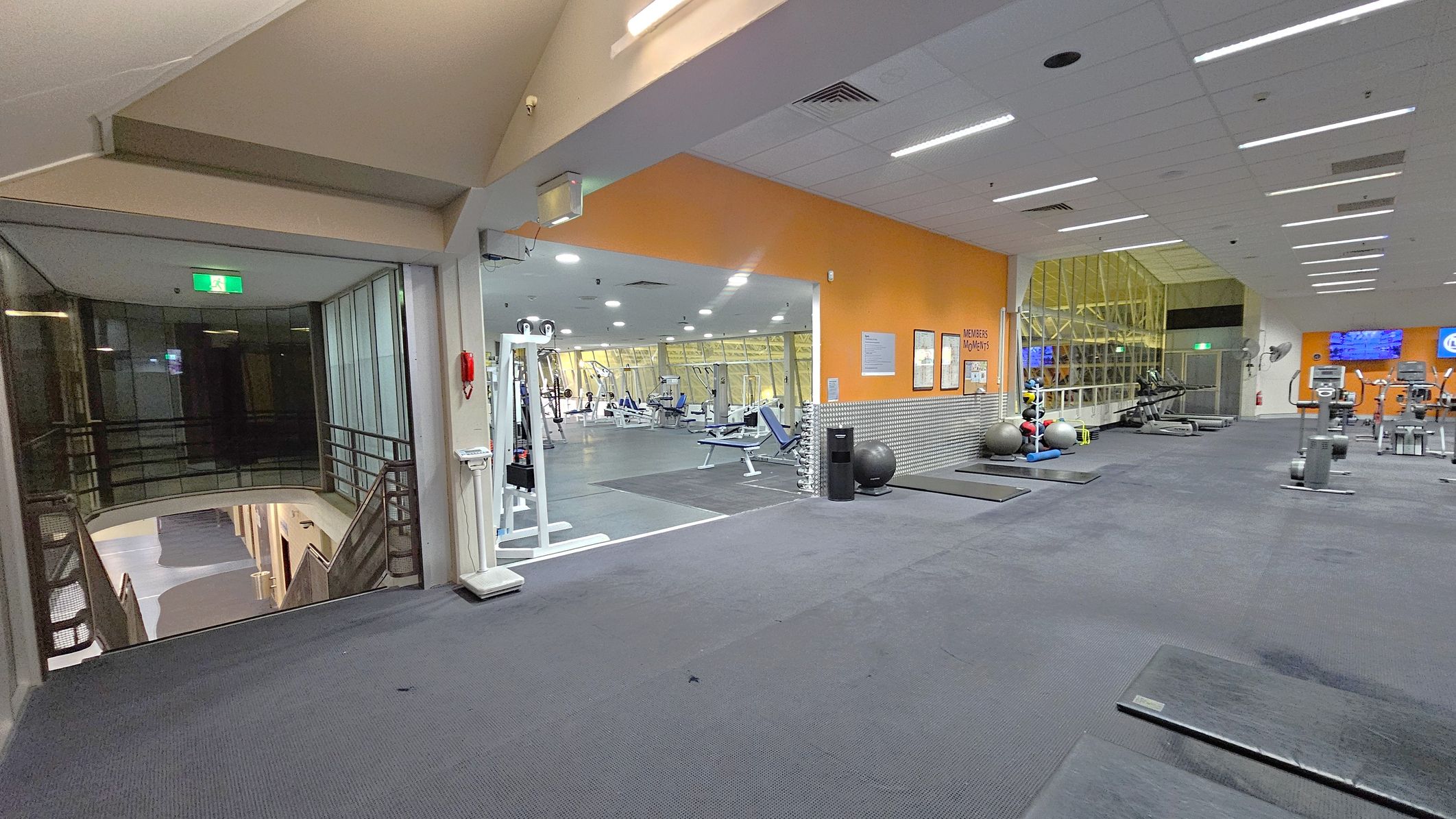 A gym interior with a grey carpeted floor and a white ceiling with recessed lighting. On the left, there's a glass-walled area with a staircase leading down. In the centre, an open space with various gym equipment, including weight machines and benches, is visible. The right side features an orange wall with posters, exercise balls, and more gym equipment like treadmills. A large window with a view of the outside is in the background.