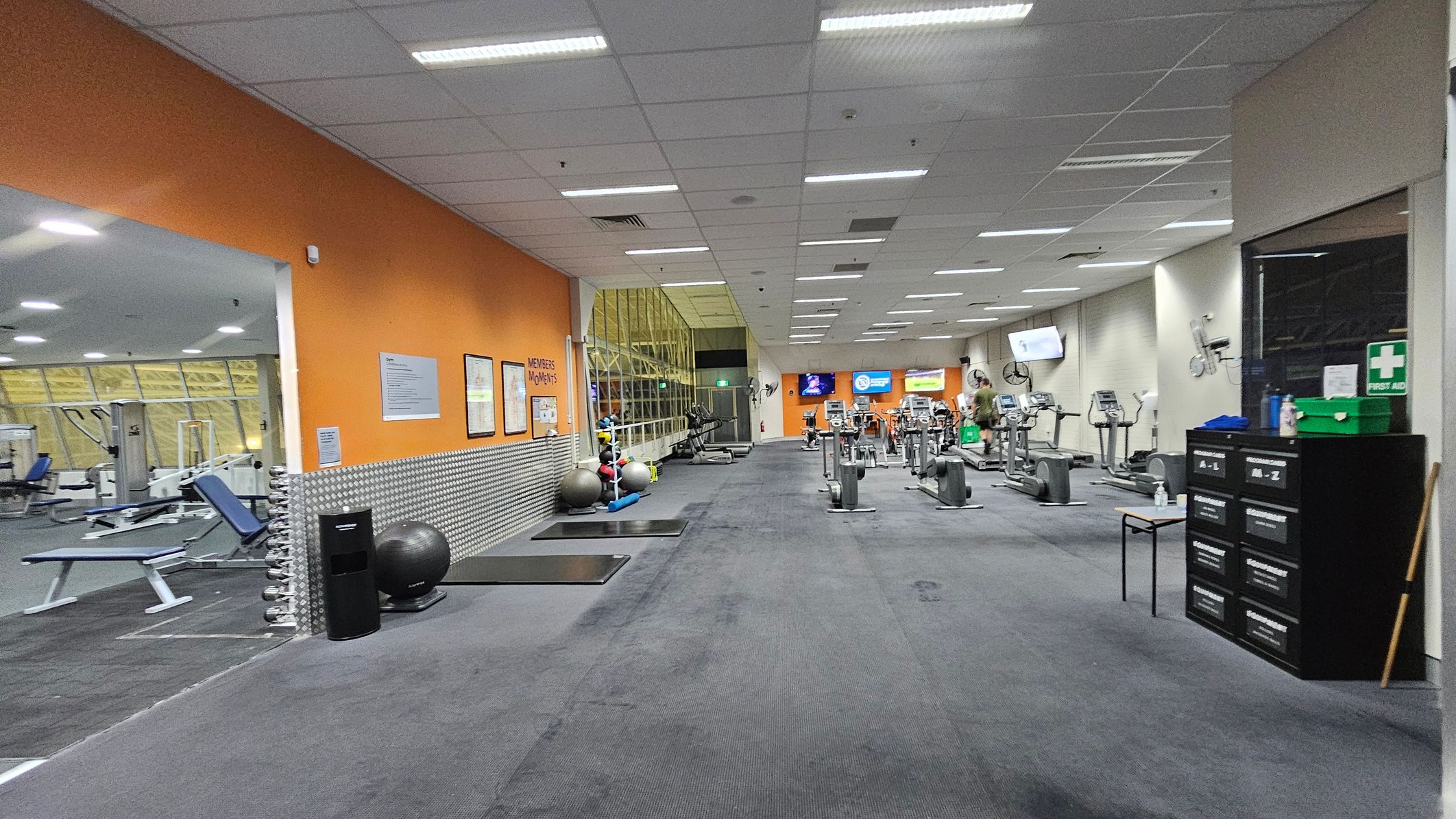 A gym interior with an orange wall on the left featuring framed posters and a rack of exercise balls. Adjacent is a room with weight benches and gym equipment. The centre has a row of exercise machines, including treadmills and stationary bikes. On the right, a black cabinet with a first aid kit on top. The ceiling is lined with fluorescent lights, and the floor is carpeted in dark grey. A large mirror spans the back wall, reflecting the gym equipment.