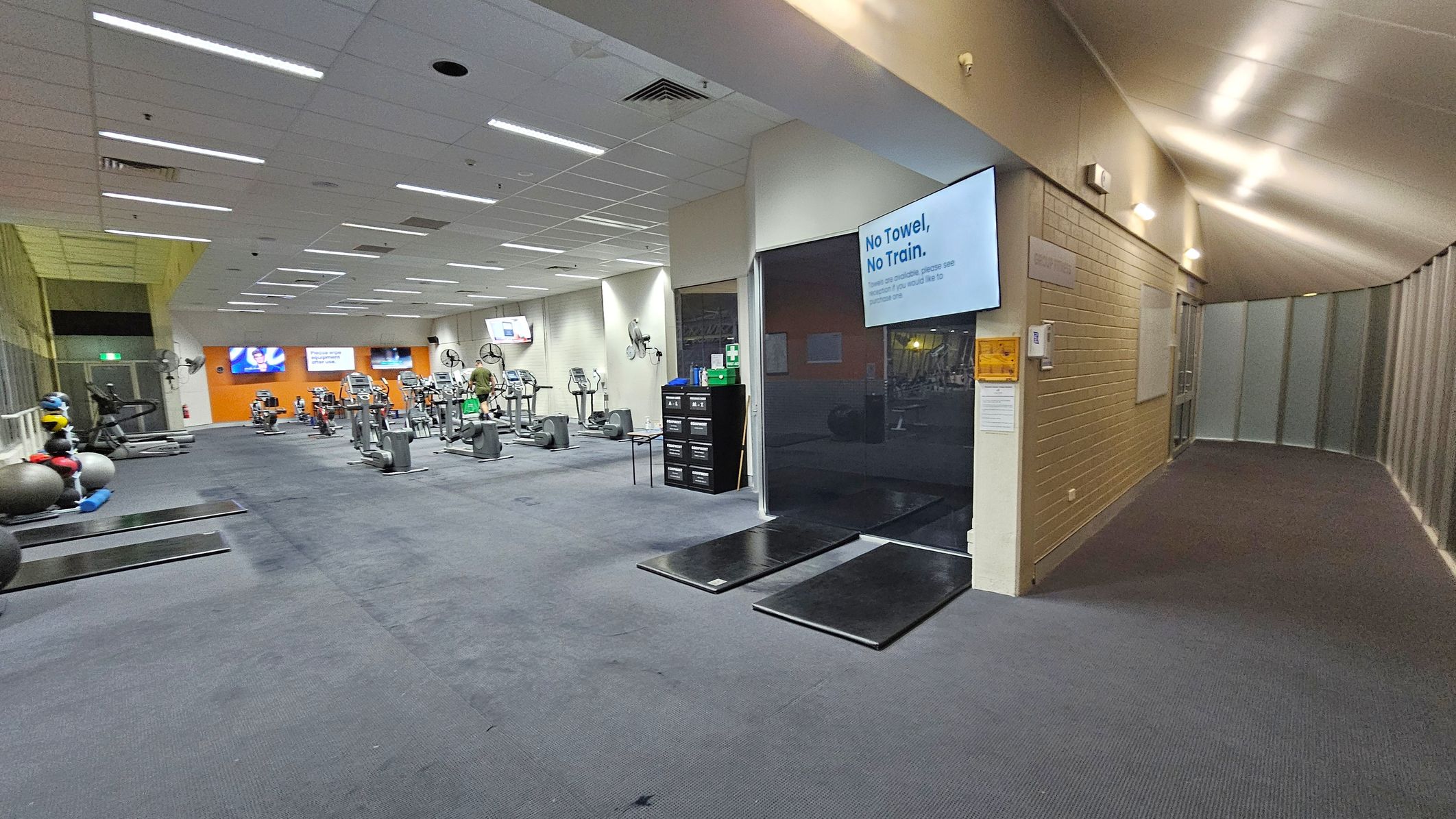 A gym interior with exercise equipment on the left, including treadmills and stationary bikes. The floor is carpeted in a dark colour. On the right, a corridor with a light-coloured wall and ceiling, illuminated by overhead lights. A sign on the wall reads "No Towel, No Train." In the background, there are orange walls with mounted screens. The ceiling has recessed lighting and ventilation panels.