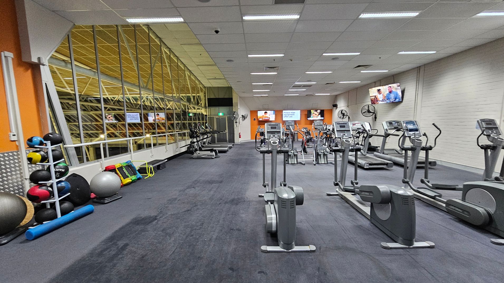 A gym with a purple carpeted floor and a white panelled ceiling. On the left, there are exercise balls and foam rollers stacked against a mirrored wall. In the centre, several elliptical machines are lined up. On the right, treadmills face a wall with mounted televisions. The back wall is orange with two screens displaying images. Ceiling lights are evenly spaced, and there are wall-mounted fans.