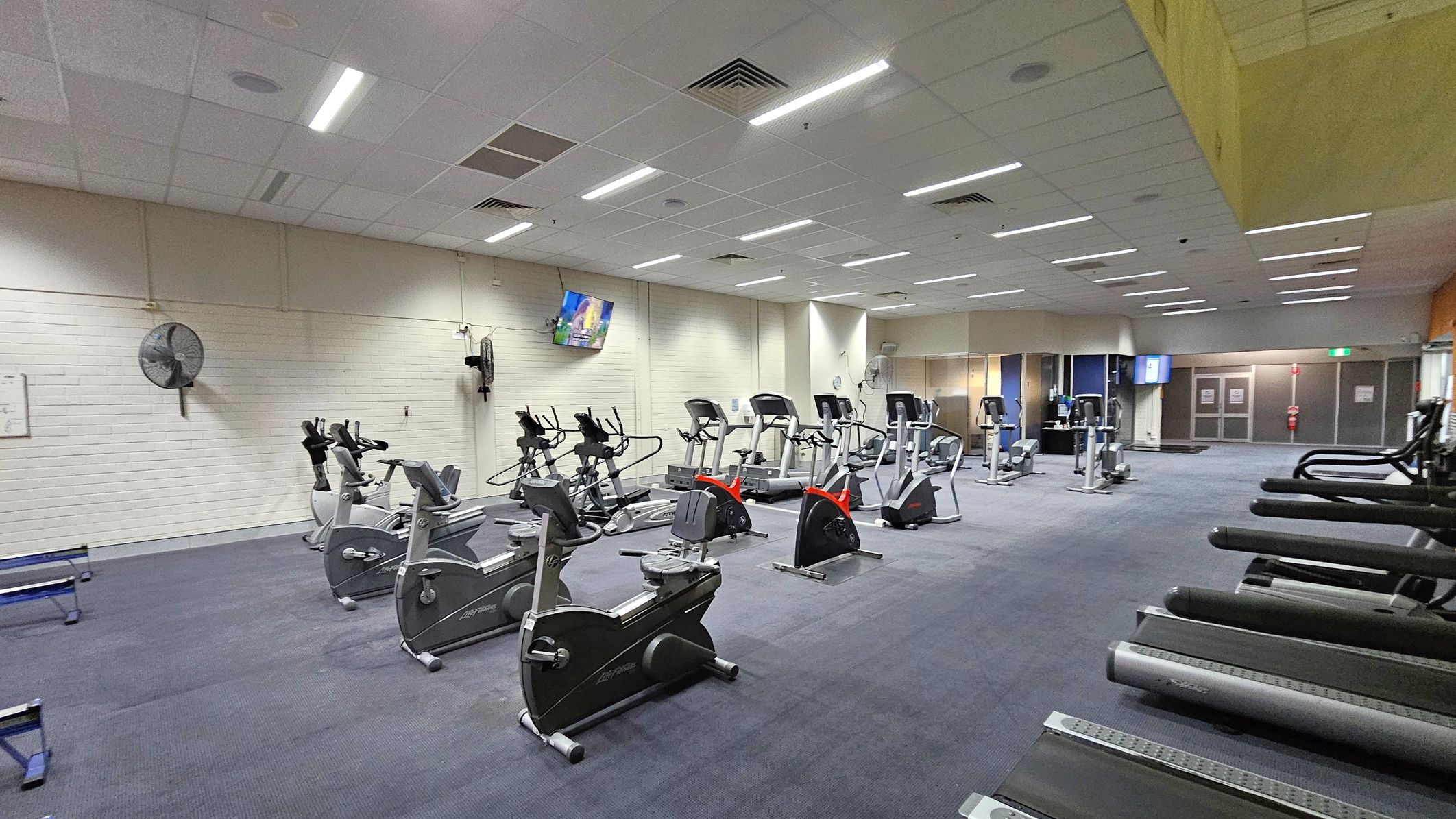 A spacious gym with a variety of exercise equipment. On the left, there are several stationary bikes and elliptical machines. In the centre, more exercise bikes and elliptical trainers are arranged in rows. On the right, a row of treadmills is visible. The floor is carpeted in a dark blue shade. The ceiling is fitted with fluorescent lights and air vents. A wall-mounted fan and a television are on the left wall, while the far end has glass doors and a fire extinguisher.