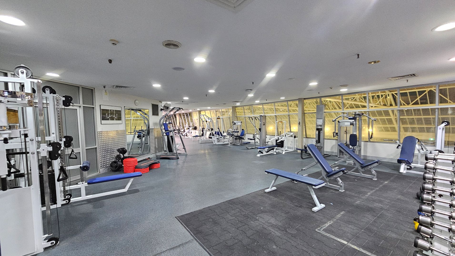 A spacious gym with various exercise equipment. On the left, there are weight machines and a stack of red and black weights. In the centre, several benches and machines are arranged, with blue padding. The right side features a rack of dumbbells. Large windows in the background allow natural light, and the ceiling is fitted with recessed lights. The floor is a mix of rubber and carpeted surfaces.