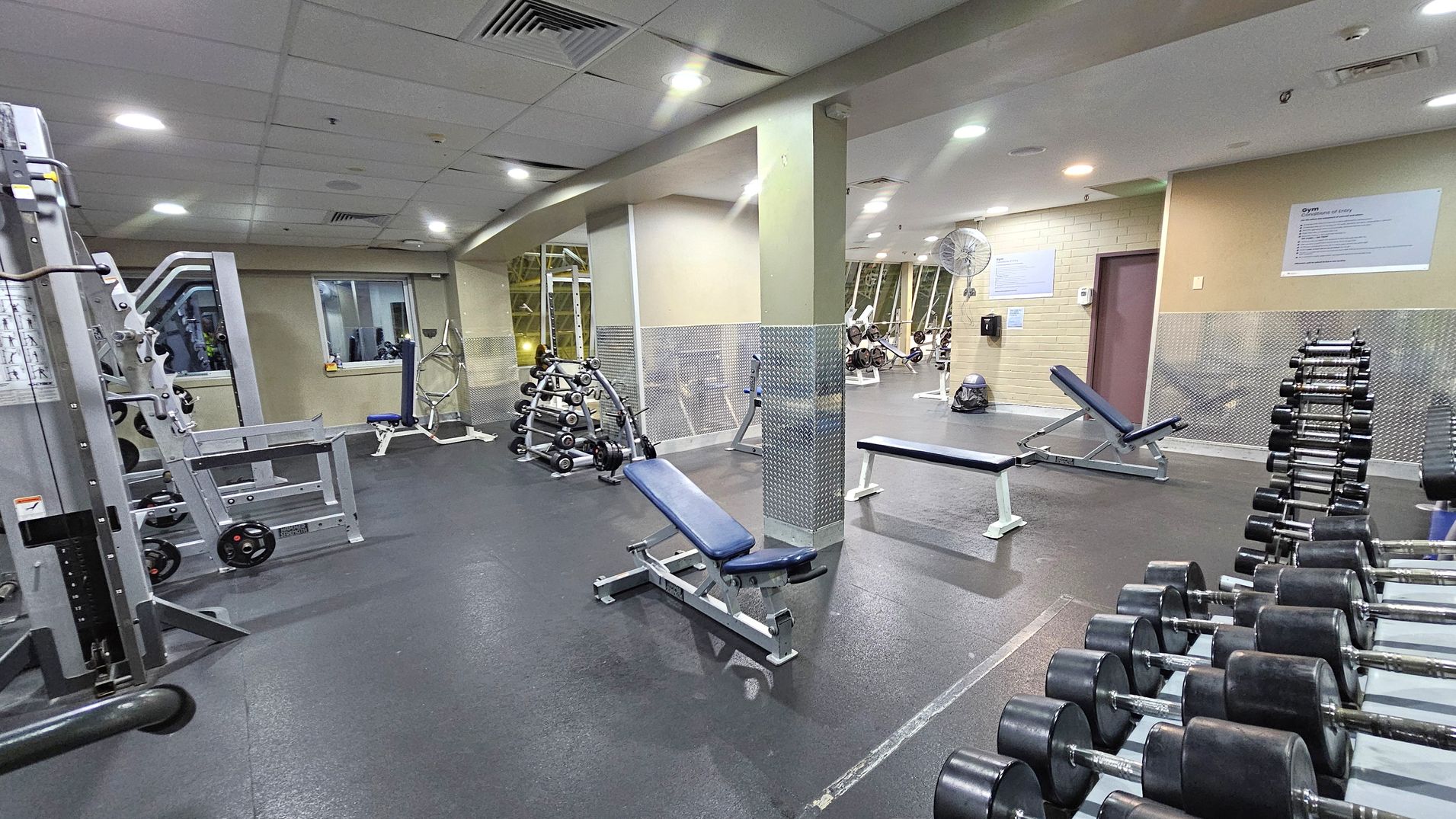 A gym with various equipment. On the left, there's a weight machine and a window. In the centre, a blue bench is positioned near a rack of dumbbells. On the right, another bench is visible with a wall-mounted fan above it. The floor is dark and rubberised, while the ceiling is fitted with recessed lights and air vents. The walls have a mix of light colours and metallic panels.