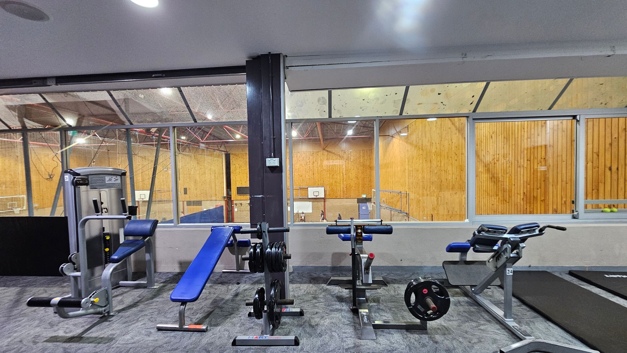 A gym area with various exercise equipment on a carpeted floor. On the left, there's a leg extension machine with a blue seat. In the centre, a weight bench with a rack of weights. On the right, a hyperextension bench and a treadmill. The background features large windows overlooking a sports hall with wooden walls and a basketball hoop. The ceiling has exposed beams and lights.