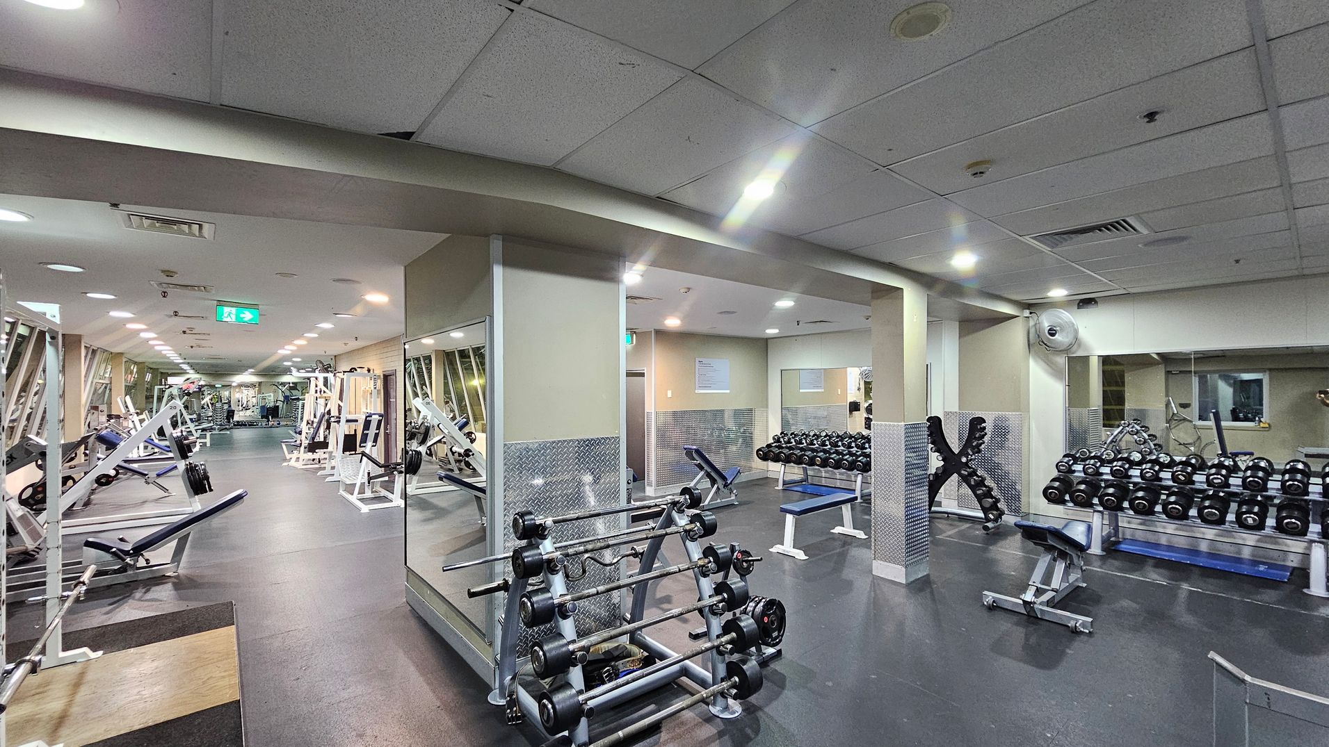 A spacious gym with a variety of equipment. On the left, there are weightlifting benches and machines with weights. In the centre, a rack holds multiple barbells. On the right, there are rows of dumbbells on racks and a bench. The floor is dark and rubberised, while the ceiling is white with recessed lighting. Mirrors line the walls, enhancing the sense of space. An emergency exit sign is visible in the background.