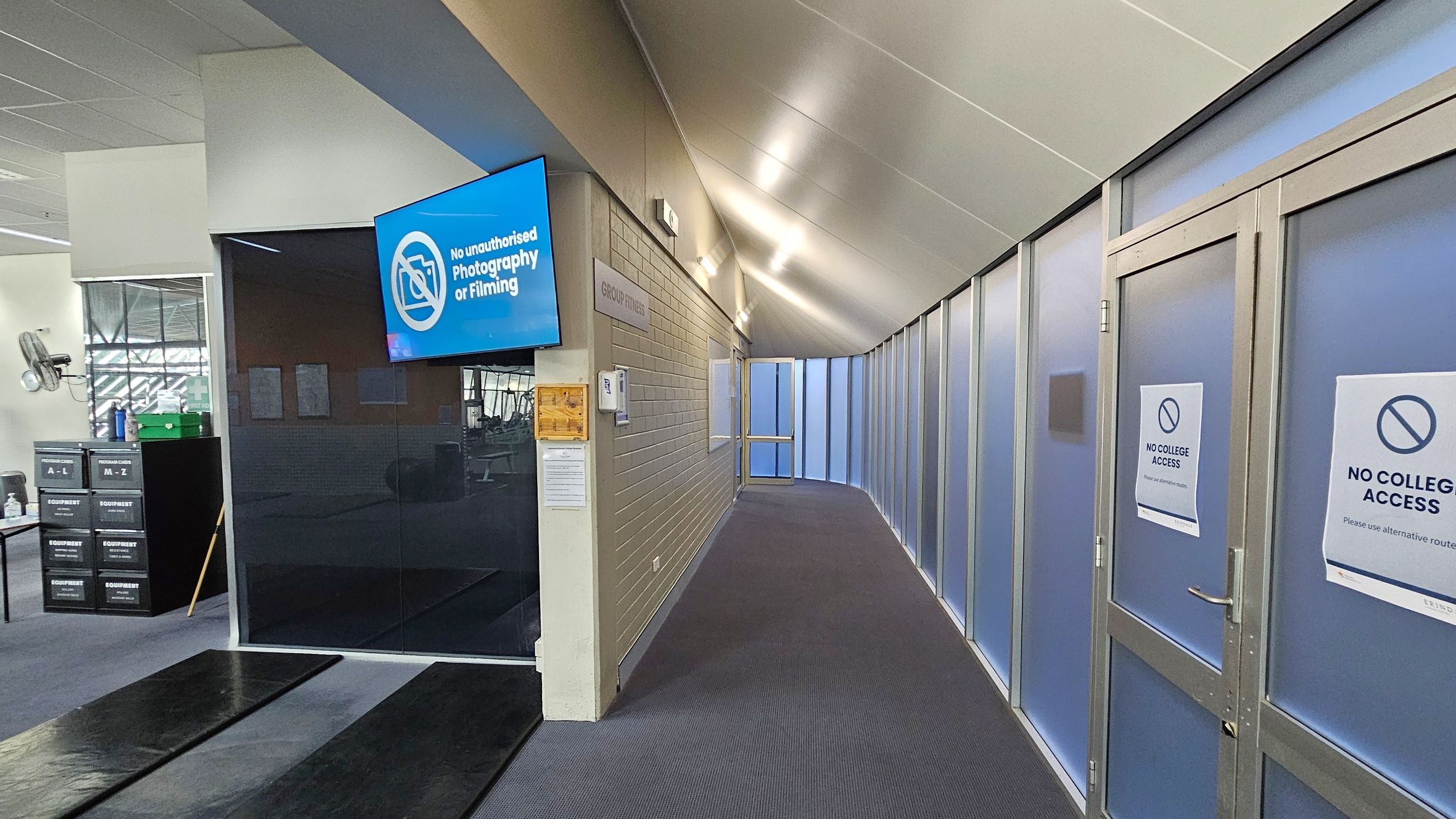 A corridor with a sloped ceiling and carpeted floor. On the left, there's a blue sign reading "No unauthorised Photography or Filming" above a dark wall. Nearby, equipment storage labelled "A-L" and "M-Z" is visible. On the right, frosted glass doors have signs stating "No College Access." The corridor leads to a door at the end, with "Group Fitness" signage on the wall. The lighting is bright, with fixtures along the ceiling.