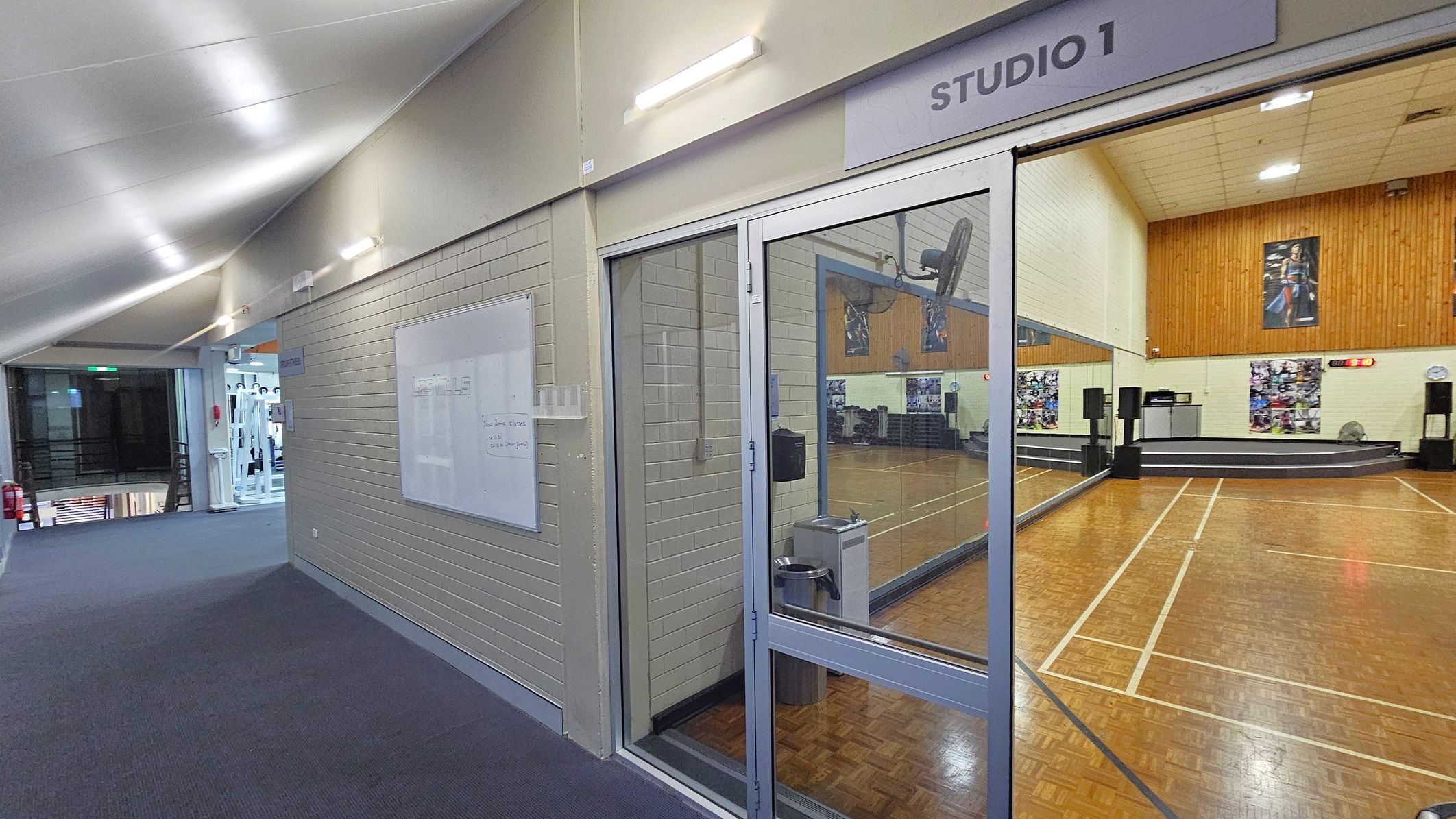 A hallway with a blue carpet leads to a glass entrance labelled "STUDIO 1" on the right. Inside the studio, there's a wooden floor with white lines, a stage with speakers, and posters on the walls. The ceiling is white with fluorescent lights. On the left, a whiteboard is mounted on a brick wall, and further down, gym equipment is visible through an open doorway. A fire extinguisher is mounted on the wall near the gym area.