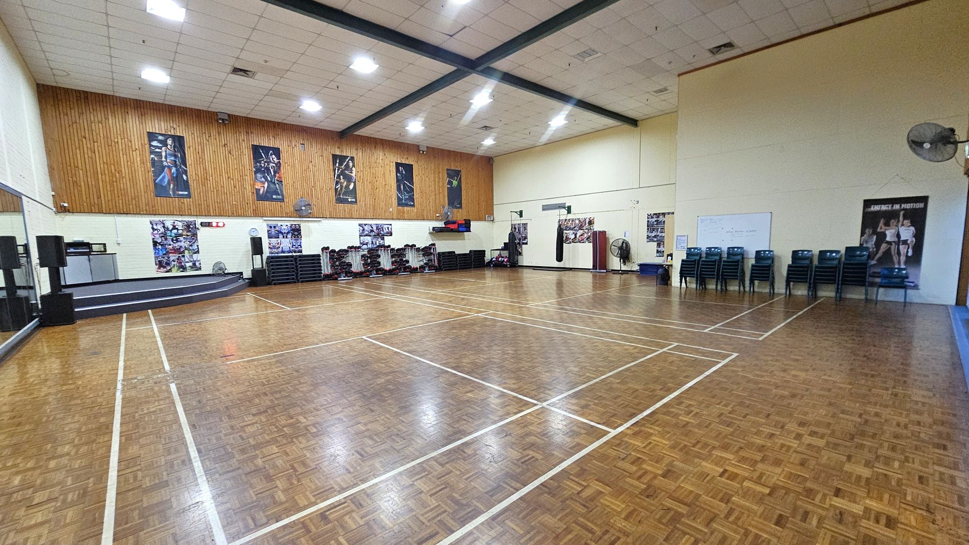 A spacious indoor gymnasium with a wooden parquet floor marked with white lines for sports. On the left, there's a stage with speakers and a large mirror. The back wall features several fitness posters and stacked exercise equipment. On the right, there are stacked chairs, a whiteboard, and a large fan. The ceiling is high with exposed beams and bright overhead lights.
