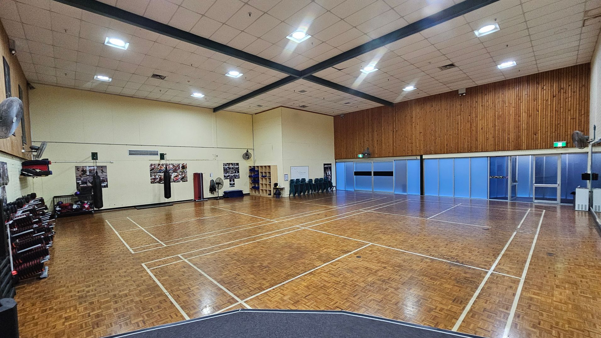 A spacious indoor gymnasium with a parquet wooden floor marked for sports. On the left, there are shelves with exercise equipment and punching bags hanging from the wall. The back wall features a wooden panel design and a stack of chairs. The right side has frosted glass doors and windows. The ceiling is high with fluorescent lights and exposed beams. Walls are painted light yellow, and there are several fans mounted on them.