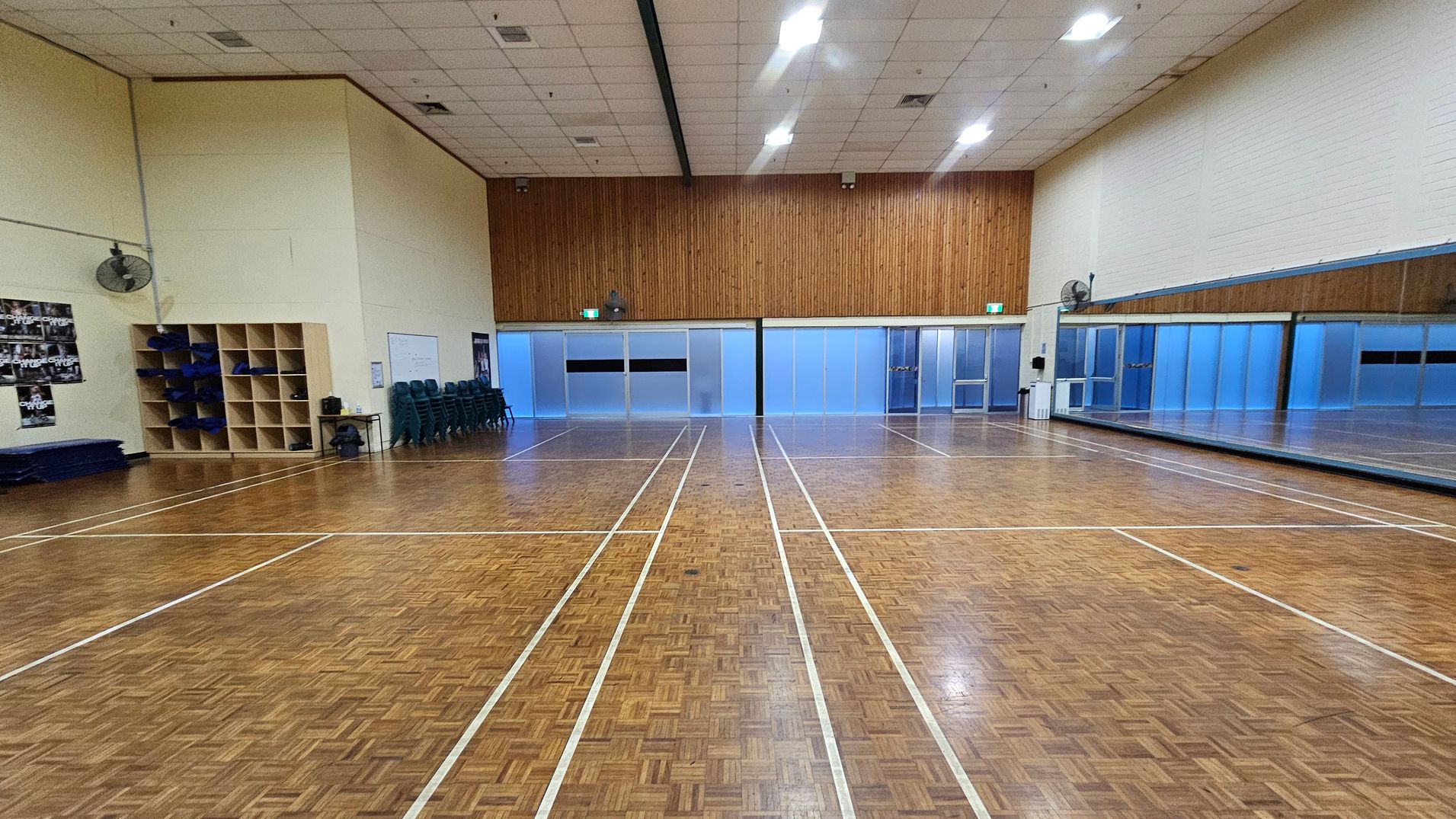 A spacious indoor sports hall with a parquet floor marked with white lines for games. On the left, there are shelves with blue mats and a stack of chairs. Posters are visible on the wall. The back wall features frosted glass panels and a wooden section above. On the right, a large mirror spans the wall, reflecting the room. The ceiling is high with fluorescent lights, and there are wall-mounted fans on both sides.