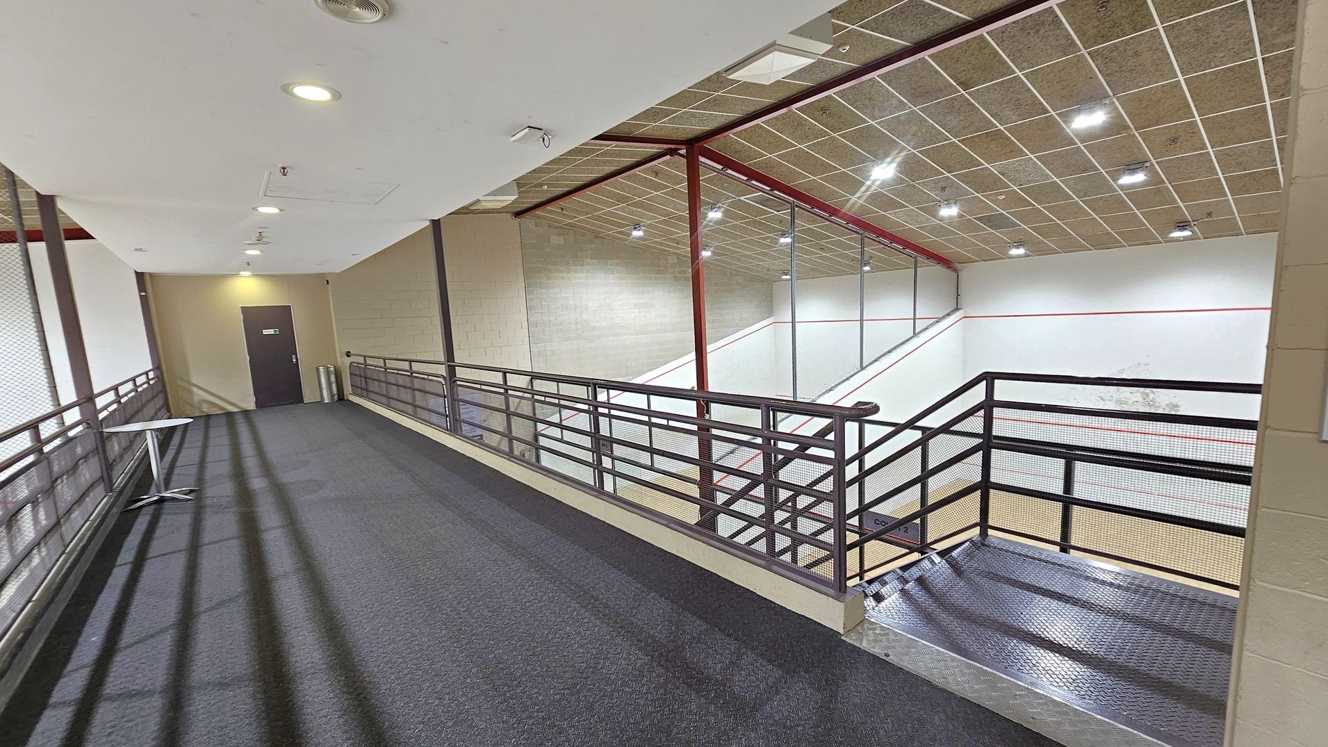 A corridor with a dark carpeted floor and a white ceiling with recessed lights. On the left, there's a small table and a door at the end of the corridor. The right side features a metal railing overlooking a squash court below, with a wooden floor and white walls marked with red lines. The ceiling above the court is tiled with multiple lights.