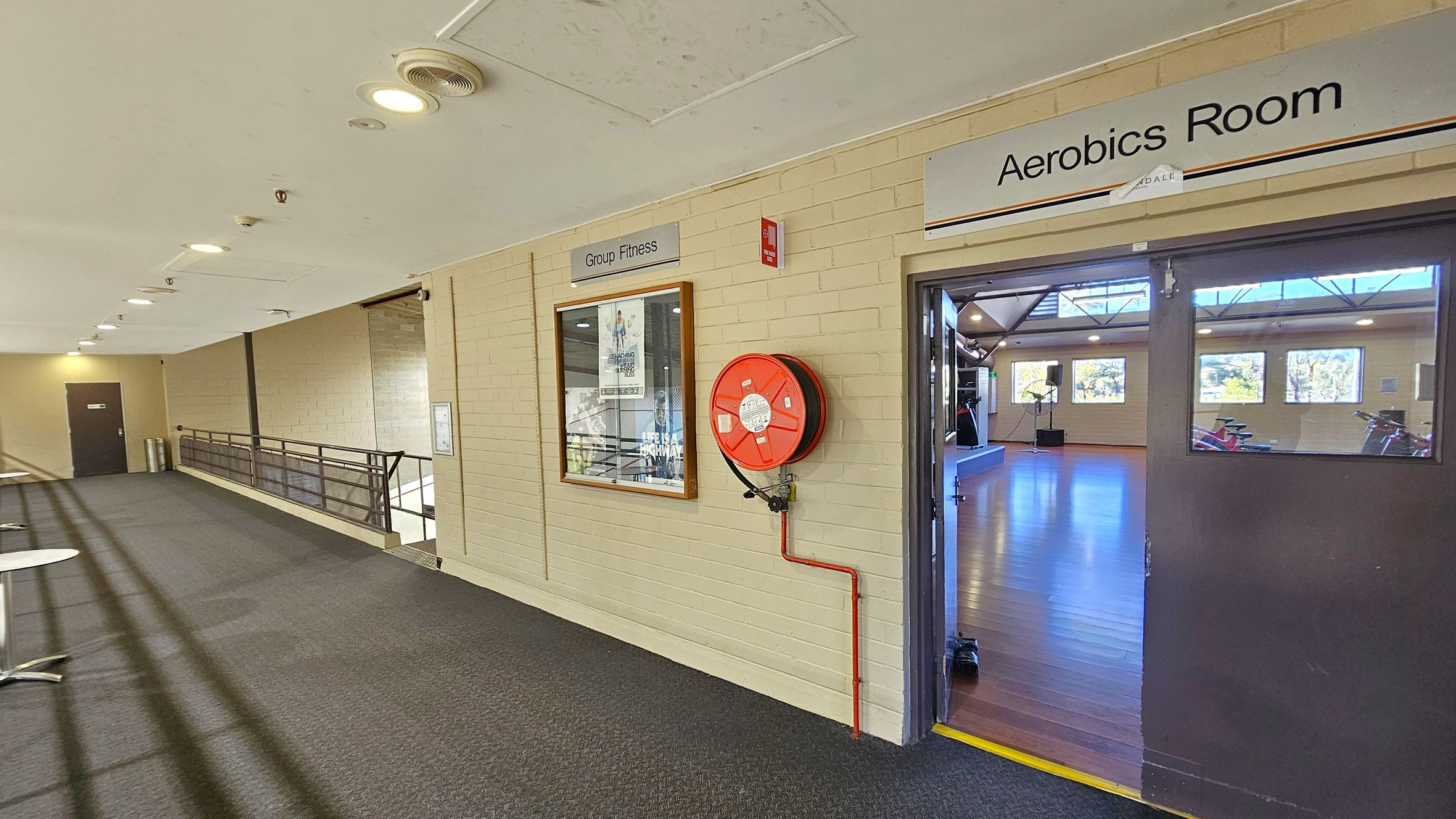 A corridor with a carpeted floor and a ceiling with recessed lighting. On the right, there's an open door leading to an "Aerobics Room" with wooden flooring and exercise equipment visible inside. A red fire hose reel is mounted on the wall next to the door. On the left, a sign reads "Group Fitness" above a framed poster. A railing runs along the corridor, and a door is visible at the far end.