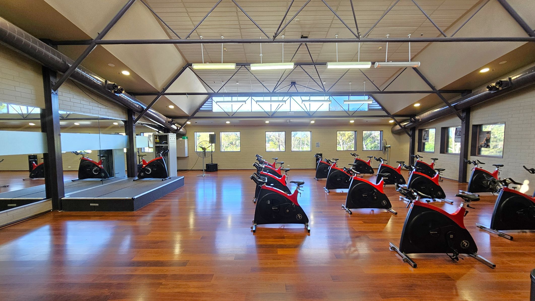 A spacious indoor cycling studio with a wooden floor. On the right, there are several stationary bikes arranged in rows. On the left, a large mirror covers the wall, reflecting the bikes and the room. The ceiling is high with exposed beams and ductwork, and fluorescent lights hang from it. The back wall has several windows allowing natural light to enter. A small platform with additional equipment is visible near the mirror.