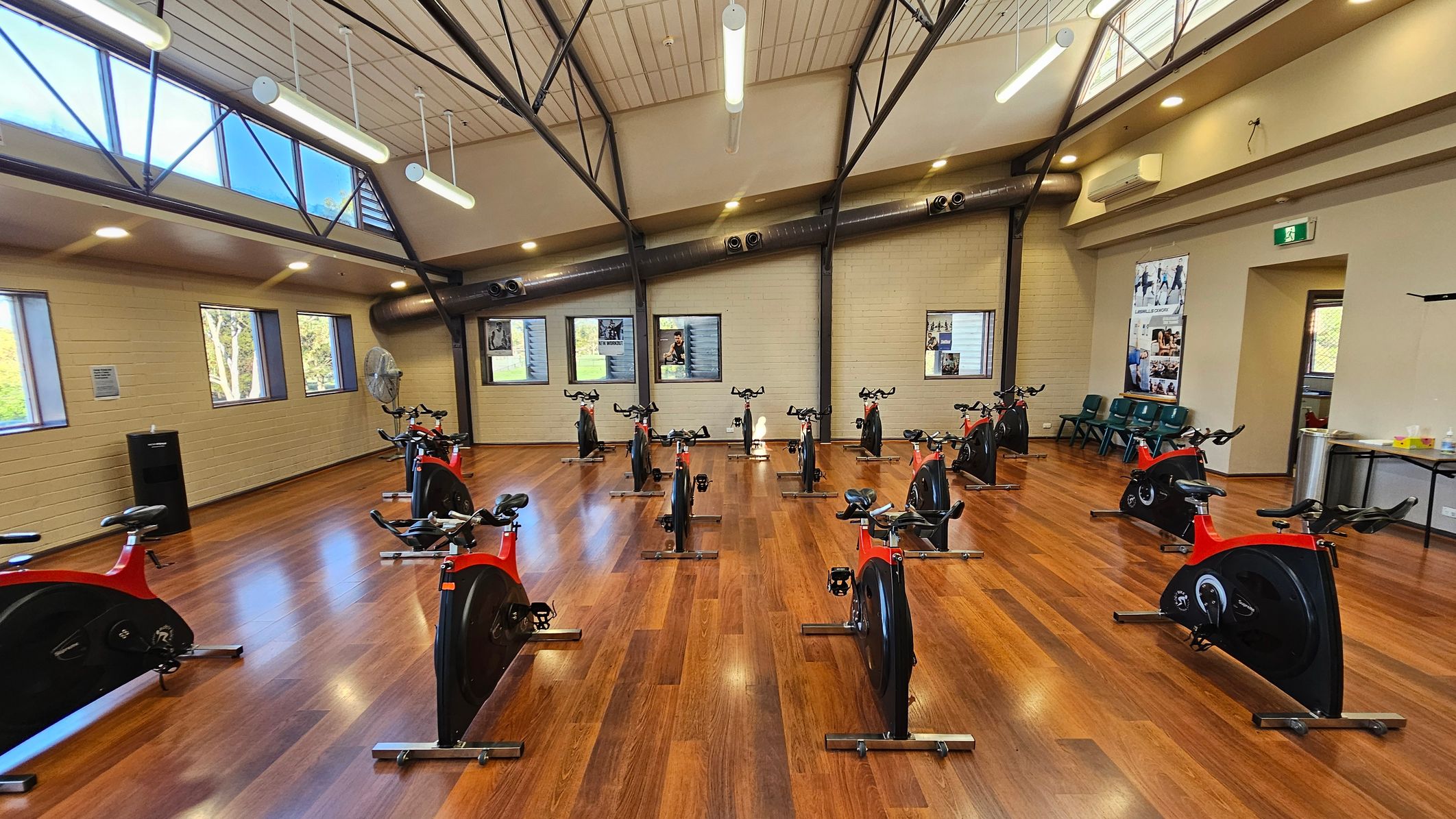 A spacious indoor cycling studio with wooden flooring and a high, sloped ceiling featuring exposed beams and fluorescent lights. The room contains several stationary bikes arranged in rows, each with a red and black design. Large windows on the left wall allow natural light to enter. The right wall has posters and a door with an exit sign above it. A few chairs and a table are positioned against the right wall. The overall atmosphere is organised and well-lit.
