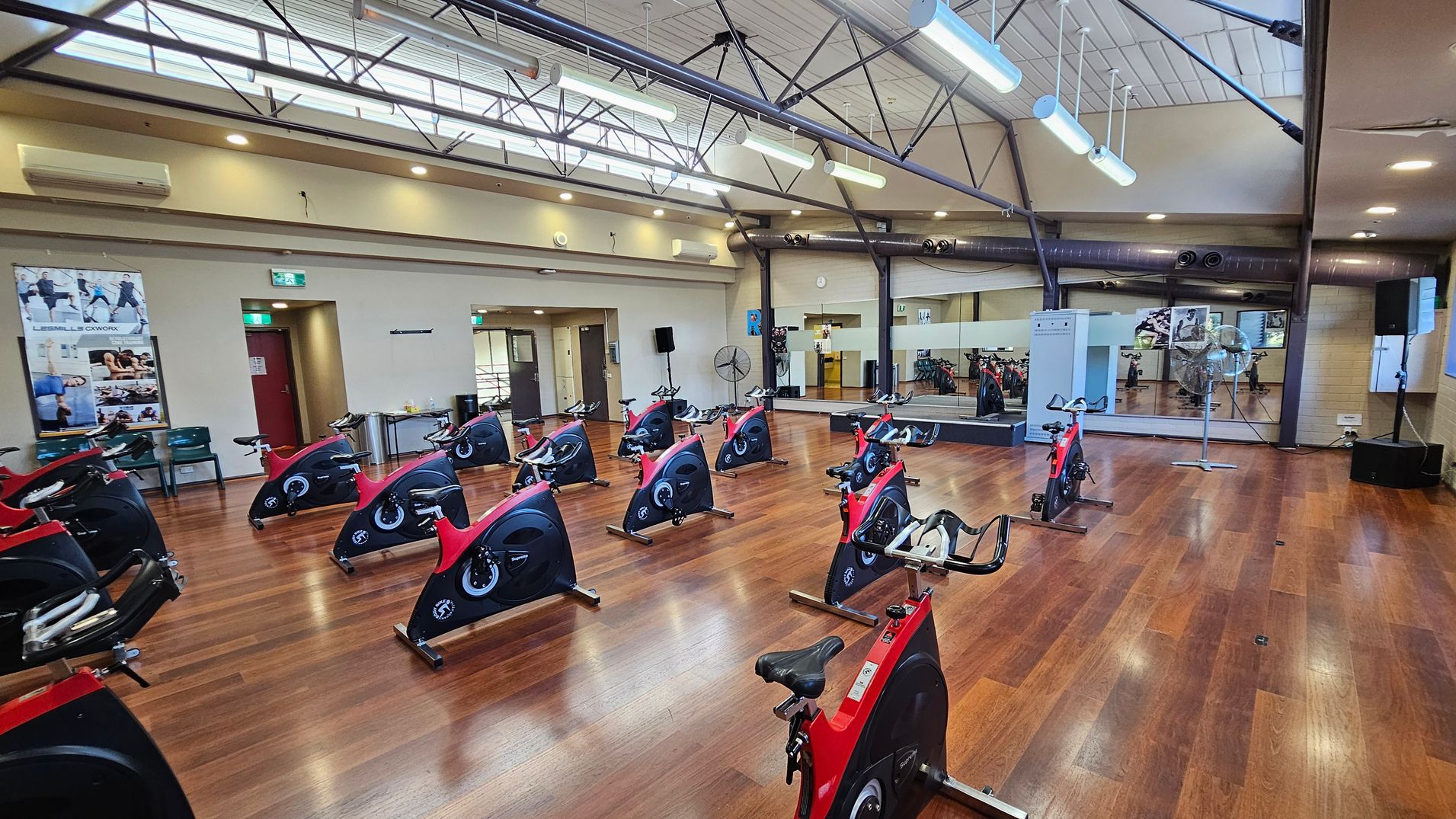 A spacious indoor cycling studio with wooden flooring. Red and black stationary bikes are arranged in rows across the room. The left wall features posters and a red door, while the right wall has large mirrors and a fan. The ceiling is high with exposed beams and fluorescent lights. A sound system is visible in the corner.