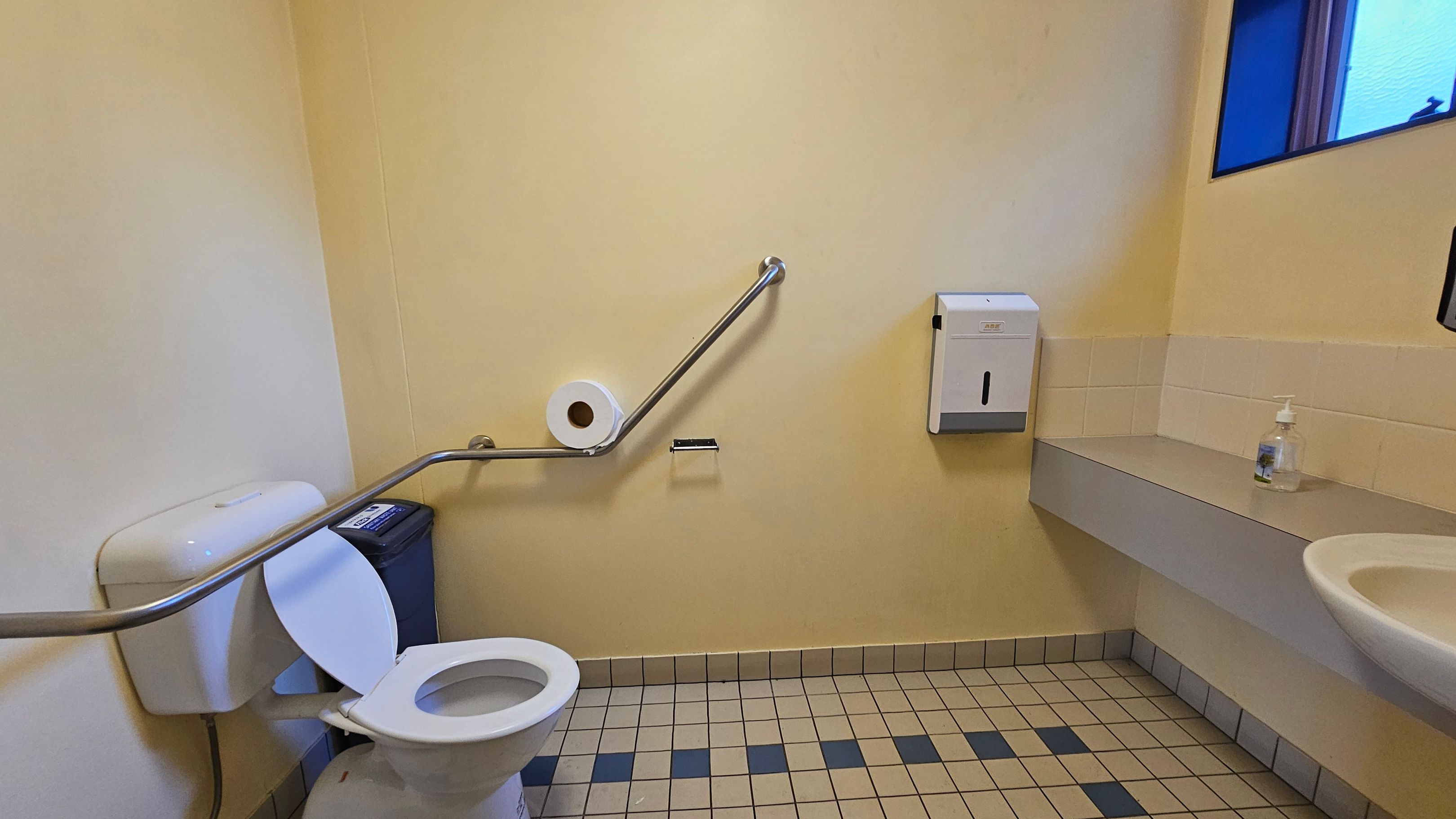 A public restroom with a beige tiled floor. On the left, there is a toilet with a raised seat and a metal grab bar. A toilet roll is mounted on the wall above the bar. On the right, a wall-mounted paper towel dispenser is above a counter with a bottle of hand sanitiser. A small window is visible at the top right. The walls are painted a light yellow.