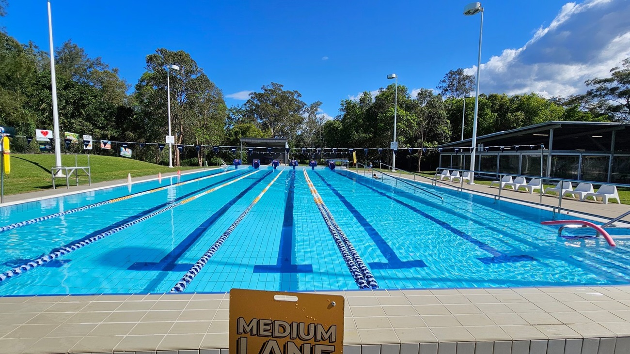 An outdoor swimming pool with multiple lanes, each separated by floating lane markers. On the left, there is a grassy area with trees and several advertising banners along a fence. In the middle, the pool has a metal handrail leading into the water. On the right, there is a concrete pool deck with white lounge chairs lined up along the side. A building with large windows is also visible on the right. Tall light poles are positioned around the pool area. An outdoor swimming pool with multiple lanes, each separated by floating lane markers. On the left, there is a grassy area with trees and several advertising banners along a fence. In the middle, the pool has a metal handrail leading into the water. On the right, there is a concrete pool deck with white lounge chairs lined up along the side. A building with large windows is also visible on the right. Tall light poles are positioned around the pool area.