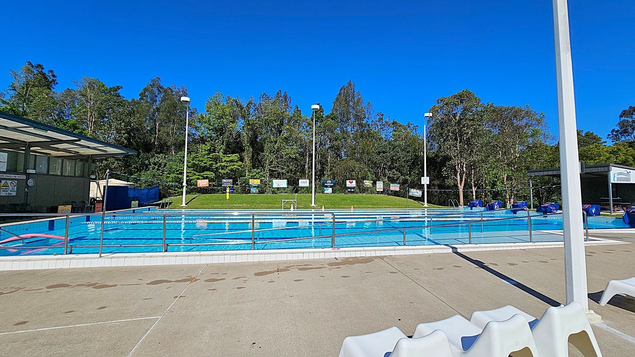 Outdoor swimming pool with clear blue water, surrounded by a metal railing. On the left, there is a covered area with signs and a small building. In the middle, the pool is divided into lanes with floating lane markers. The background features a grassy hill with various signs and tall trees. On the right, there are several blue pool covers and another small structure. The floor around the pool is concrete with some water spots. Outdoor swimming pool with clear blue water, surrounded by a metal railing. On the left, there is a covered area with signs and a small building. In the middle, the pool is divided into lanes with floating lane markers. The background features a grassy hill with various signs and tall trees. On the right, there are several blue pool covers and another small structure. The floor around the pool is concrete with some water spots.