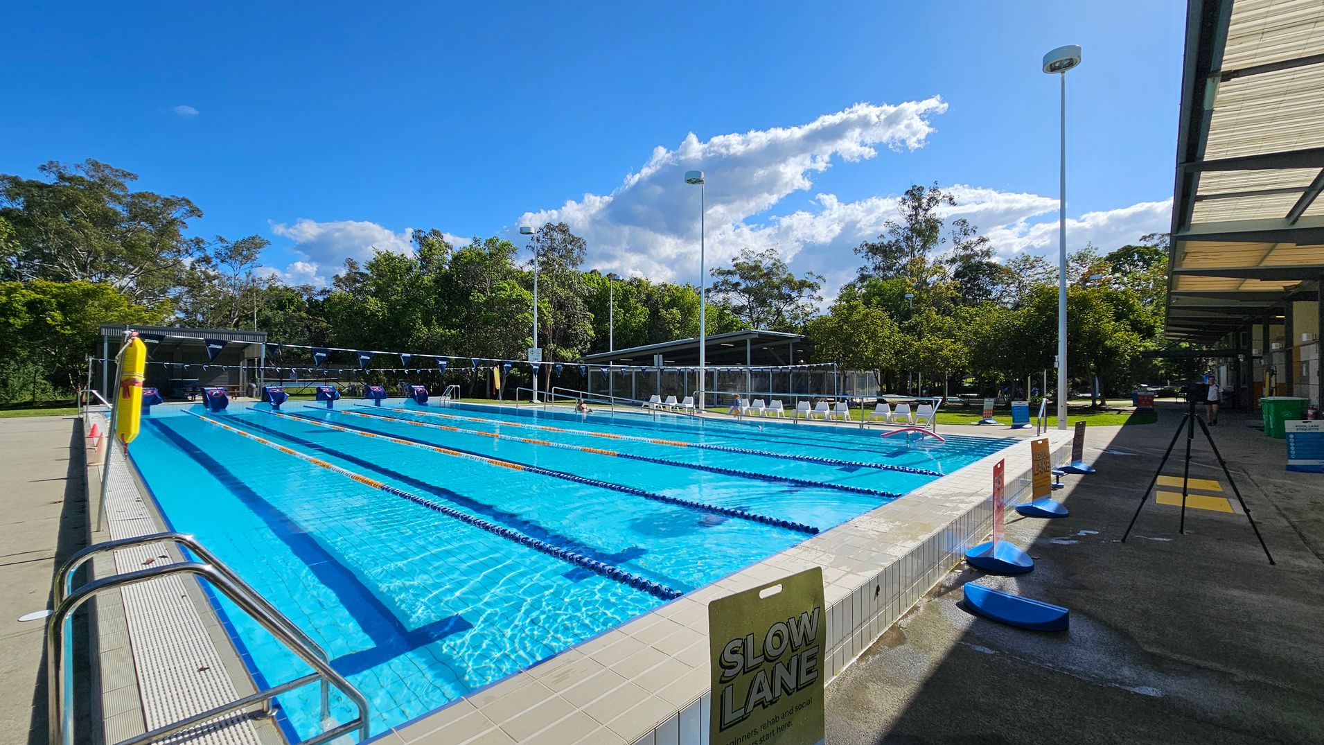 An outdoor swimming pool with multiple lanes, each marked by blue and yellow lane dividers. On the left, there is a metal handrail and a sign indicating the "Slow Lane." The pool deck is tiled. On the right, there is a covered walkway with a tripod and various signs. In the background, there are trees and a shaded seating area with white chairs. Tall light poles are positioned around the pool area. The floor is concrete, and the ceiling of the walkway is made of translucent panels. An outdoor swimming pool with multiple lanes, each marked by blue and yellow lane dividers. On the left, there is a metal handrail and a sign indicating the "Slow Lane." The pool deck is tiled. On the right, there is a covered walkway with a tripod and various signs. In the background, there are trees and a shaded seating area with white chairs. Tall light poles are positioned around the pool area. The floor is concrete, and the ceiling of the walkway is made of translucent panels.