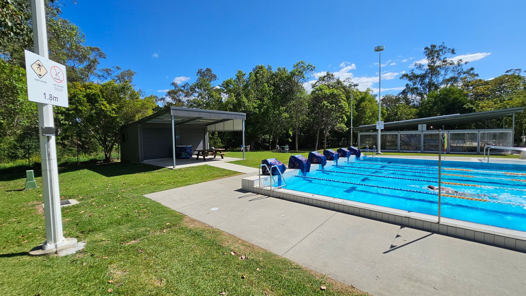 Outdoor swimming pool with lanes on the right, covered by blue lane markers. A swimmer is in one of the lanes. On the left, a sign on a pole indicates "Deep Water" and "No Diving" with a depth of 1.8m. Behind the sign, there is a shaded seating area with picnic tables. The background features green grass, trees, and a fence. The foreground has a concrete pool deck and grassy area. Outdoor swimming pool with lanes on the right, covered by blue lane markers. A swimmer is in one of the lanes. On the left, a sign on a pole indicates "Deep Water" and "No Diving" with a depth of 1.8m. Behind the sign, there is a shaded seating area with picnic tables. The background features green grass, trees, and a fence. The foreground has a concrete pool deck and grassy area.