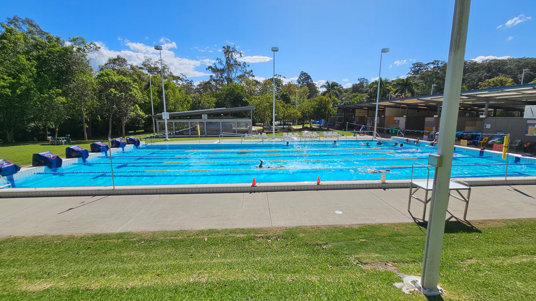 An outdoor swimming pool with multiple lanes, each marked by lane ropes. Swimmers are in the pool, and starting blocks are positioned at one end. On the left, there is a grassy area with trees and some chairs. On the right, there is a covered area with seating and various facilities. The pool deck is concrete, and there are a few orange cones placed along the edge. A tall light pole is in the foreground on the right side. An outdoor swimming pool with multiple lanes, each marked by lane ropes. Swimmers are in the pool, and starting blocks are positioned at one end. On the left, there is a grassy area with trees and some chairs. On the right, there is a covered area with seating and various facilities. The pool deck is concrete, and there are a few orange cones placed along the edge. A tall light pole is in the foreground on the right side.