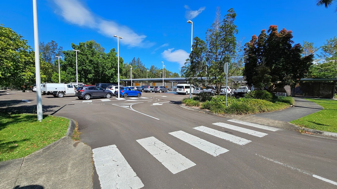A car park with several vehicles, including a white utility vehicle and a blue car, is in the middle. The foreground has a zebra crossing on an asphalt road. On the left, there is a grassy area with a tree and a tall streetlight. On the right, there are more trees, a footpath, and a grassy patch. In the background, there is a low building with a covered walkway and more parked cars. Tall streetlights are scattered throughout the car park. A car park with several vehicles, including a white utility vehicle and a blue car, is in the middle. The foreground has a zebra crossing on an asphalt road. On the left, there is a grassy area with a tree and a tall streetlight. On the right, there are more trees, a footpath, and a grassy patch. In the background, there is a low building with a covered walkway and more parked cars. Tall streetlights are scattered throughout the car park.