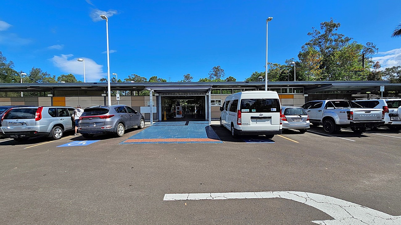 A car park with several vehicles, including a silver SUV, a grey SUV, a white van, and a white pickup truck. The car park has designated disabled parking spaces marked in blue. In the centre, there is a covered entrance to a building with a blue-tiled walkway leading to it. The building has a modern design with a mix of grey and beige panels. Trees and greenery are visible in the background. The ground is asphalt, and there are tall streetlights on either side. A car park with several vehicles, including a silver SUV, a grey SUV, a white van, and a white pickup truck. The car park has designated disabled parking spaces marked in blue. In the centre, there is a covered entrance to a building with a blue-tiled walkway leading to it. The building has a modern design with a mix of grey and beige panels. Trees and greenery are visible in the background. The ground is asphalt, and there are tall streetlights on either side.