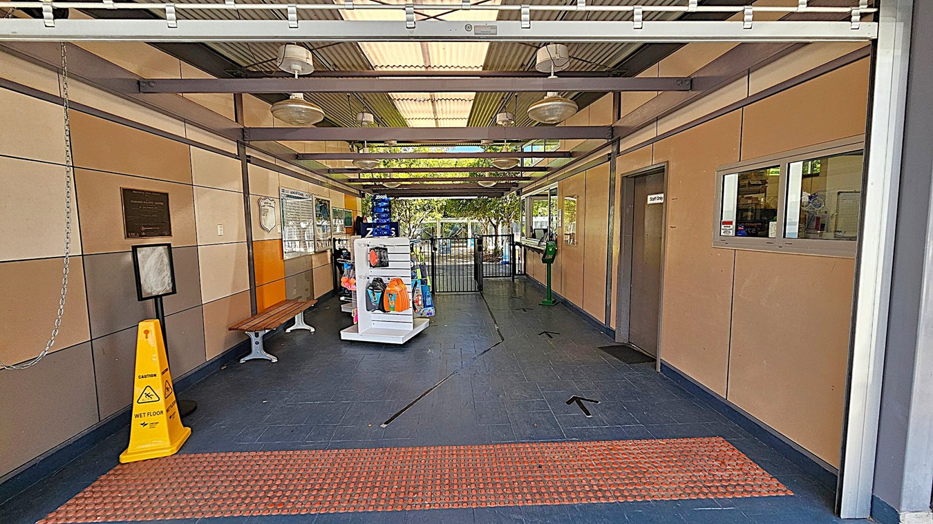 A corridor with a dark tiled floor and a ceiling with exposed beams and hanging lights. On the left, there is a yellow caution sign, a bench, and a wall with plaques. In the middle, a white display stand holds various items, and a gate is visible further back. On the right, there is a "Staff Only" door and a window with a counter. The background shows trees and outdoor light coming through the gate. A corridor with a dark tiled floor and a ceiling with exposed beams and hanging lights. On the left, there is a yellow caution sign, a bench, and a wall with plaques. In the middle, a white display stand holds various items, and a gate is visible further back. On the right, there is a "Staff Only" door and a window with a counter. The background shows trees and outdoor light coming through the gate.
