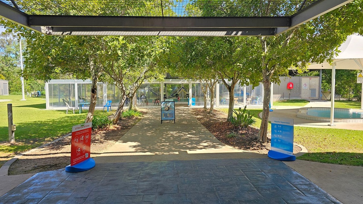 A paved pathway leads through a garden area with trees on both sides. In the foreground, there are two signs, one red and one blue, providing information. To the left, there is a grassy area with a transparent structure containing chairs. To the right, there is a small circular pool with a white canopy overhead. In the background, a glass building with people inside is visible. The floor is paved, and the ceiling is a metal grid structure. A paved pathway leads through a garden area with trees on both sides. In the foreground, there are two signs, one red and one blue, providing information. To the left, there is a grassy area with a transparent structure containing chairs. To the right, there is a small circular pool with a white canopy overhead. In the background, a glass building with people inside is visible. The floor is paved, and the ceiling is a metal grid structure.