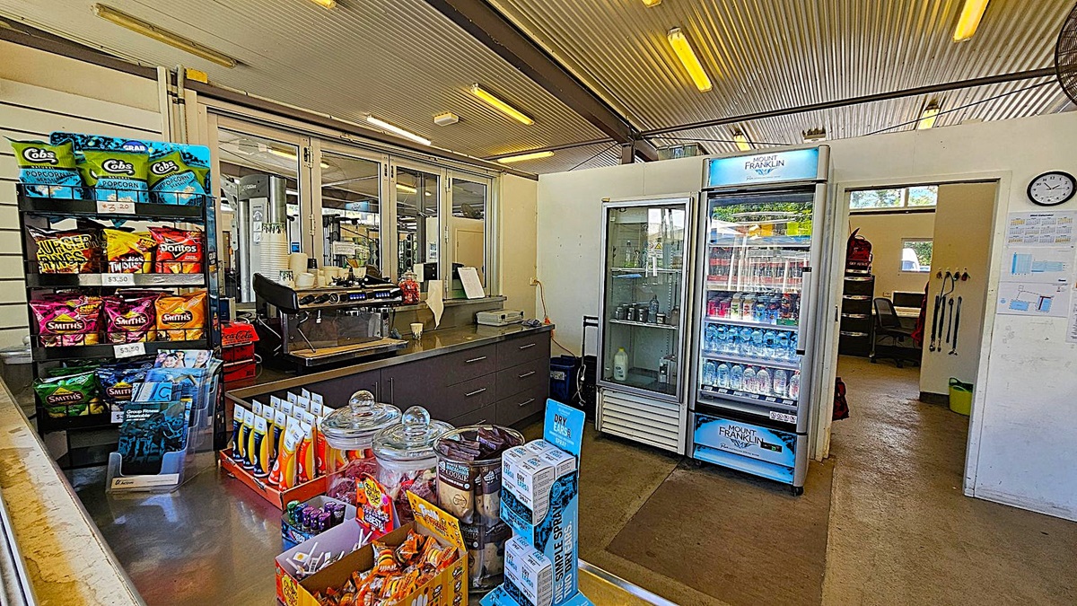 A small shop interior with a corrugated metal ceiling and concrete floor. On the left, there is a display rack with various snack packets, including popcorn and crisps. In the middle, there is a counter with a coffee machine, jars of sweets, and other small items. On the right, there is a refrigerator stocked with bottled water and other drinks. Behind the refrigerator, there is an open doorway leading to a small office area with a desk and chair. A small shop interior with a corrugated metal ceiling and concrete floor. On the left, there is a display rack with various snack packets, including popcorn and crisps. In the middle, there is a counter with a coffee machine, jars of sweets, and other small items. On the right, there is a refrigerator stocked with bottled water and other drinks. Behind the refrigerator, there is an open doorway leading to a small office area with a desk and chair.