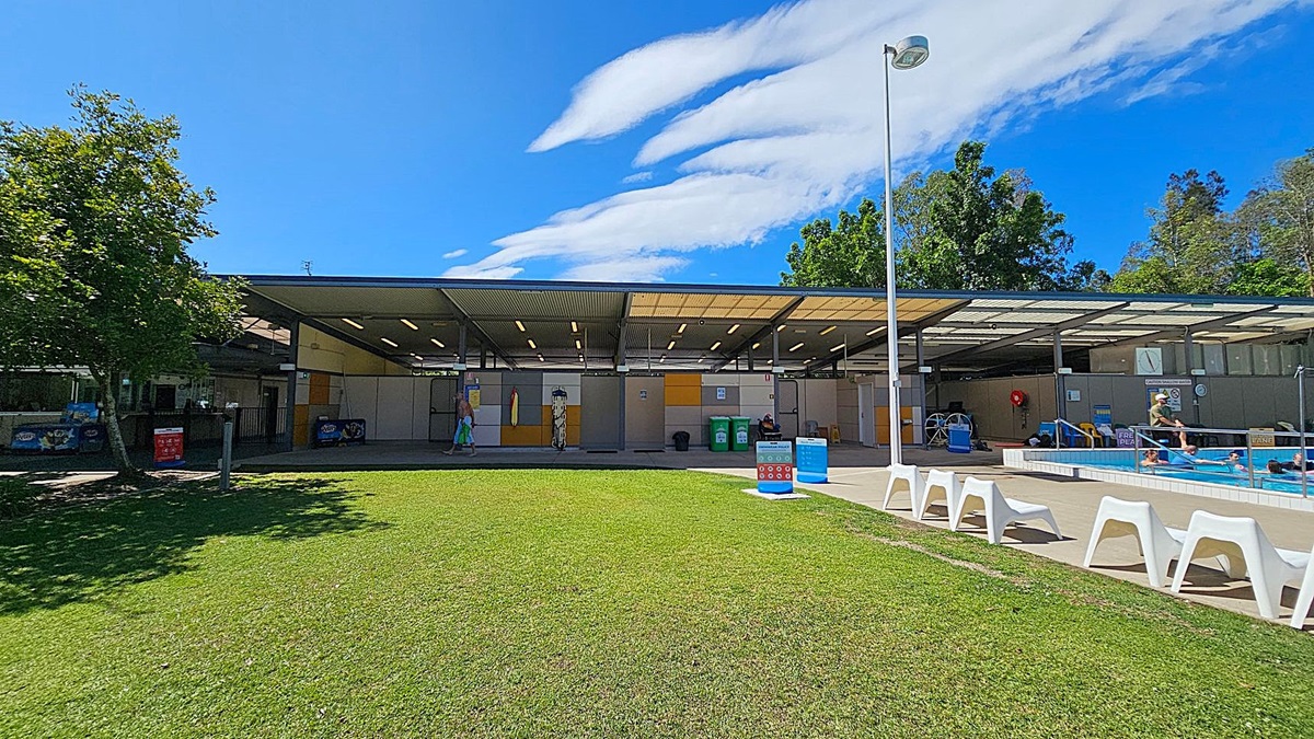 A grassy area in the foreground leads to a building with a corrugated metal roof in the background. On the left, there is a tree providing some shade. The building has open sections with people visible inside, and there are various signs and bins near the entrance. On the right, there is a swimming pool with people in it, and a row of white plastic lounge chairs along the poolside. A tall lamp post stands near the pool area. Trees are visible behind the building. A grassy area in the foreground leads to a building with a corrugated metal roof in the background. On the left, there is a tree providing some shade. The building has open sections with people visible inside, and there are various signs and bins near the entrance. On the right, there is a swimming pool with people in it, and a row of white plastic lounge chairs along the poolside. A tall lamp post stands near the pool area. Trees are visible behind the building.