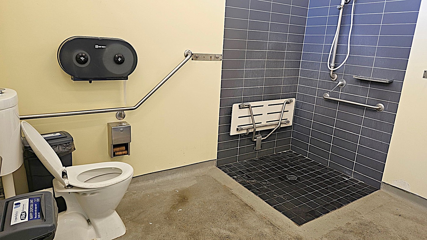 A bathroom with a toilet on the left, featuring a raised lid and a sanitary bin beside it. Above the toilet is a dual toilet paper dispenser and a grab bar. To the right is a shower area with dark grey tiles, a wall-mounted foldable shower seat, and multiple grab bars. The floor is a mix of concrete and tiled surfaces. The walls are painted light yellow. A bathroom with a toilet on the left, featuring a raised lid and a sanitary bin beside it. Above the toilet is a dual toilet paper dispenser and a grab bar. To the right is a shower area with dark grey tiles, a wall-mounted foldable shower seat, and multiple grab bars. The floor is a mix of concrete and tiled surfaces. The walls are painted light yellow.