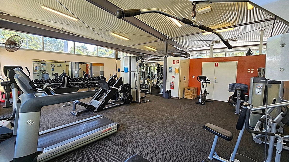 A gym with various exercise equipment. On the left, there are treadmills and a fan mounted on the wall. In the middle, there are rows of dumbbells and a leg press machine. On the right, there are stationary bikes, a fire extinguisher, and a door with an orange wall behind it. The ceiling is corrugated metal with fluorescent lights, and the floor is carpeted. Large windows near the ceiling let in natural light. A gym with various exercise equipment. On the left, there are treadmills and a fan mounted on the wall. In the middle, there are rows of dumbbells and a leg press machine. On the right, there are stationary bikes, a fire extinguisher, and a door with an orange wall behind it. The ceiling is corrugated metal with fluorescent lights, and the floor is carpeted. Large windows near the ceiling let in natural light.