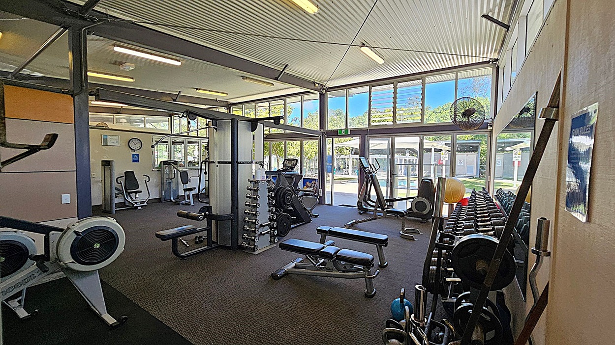 A gym with various exercise equipment. On the left, there are rowing machines and a seated exercise machine. In the middle, there is a multi-functional weight machine, benches, and a rack of dumbbells. On the right, there are more weights, exercise balls, and a large mirror. The background features large windows and glass doors, allowing natural light to enter. The ceiling is corrugated with fluorescent lights, and the floor is carpeted. A gym with various exercise equipment. On the left, there are rowing machines and a seated exercise machine. In the middle, there is a multi-functional weight machine, benches, and a rack of dumbbells. On the right, there are more weights, exercise balls, and a large mirror. The background features large windows and glass doors, allowing natural light to enter. The ceiling is corrugated with fluorescent lights, and the floor is carpeted.