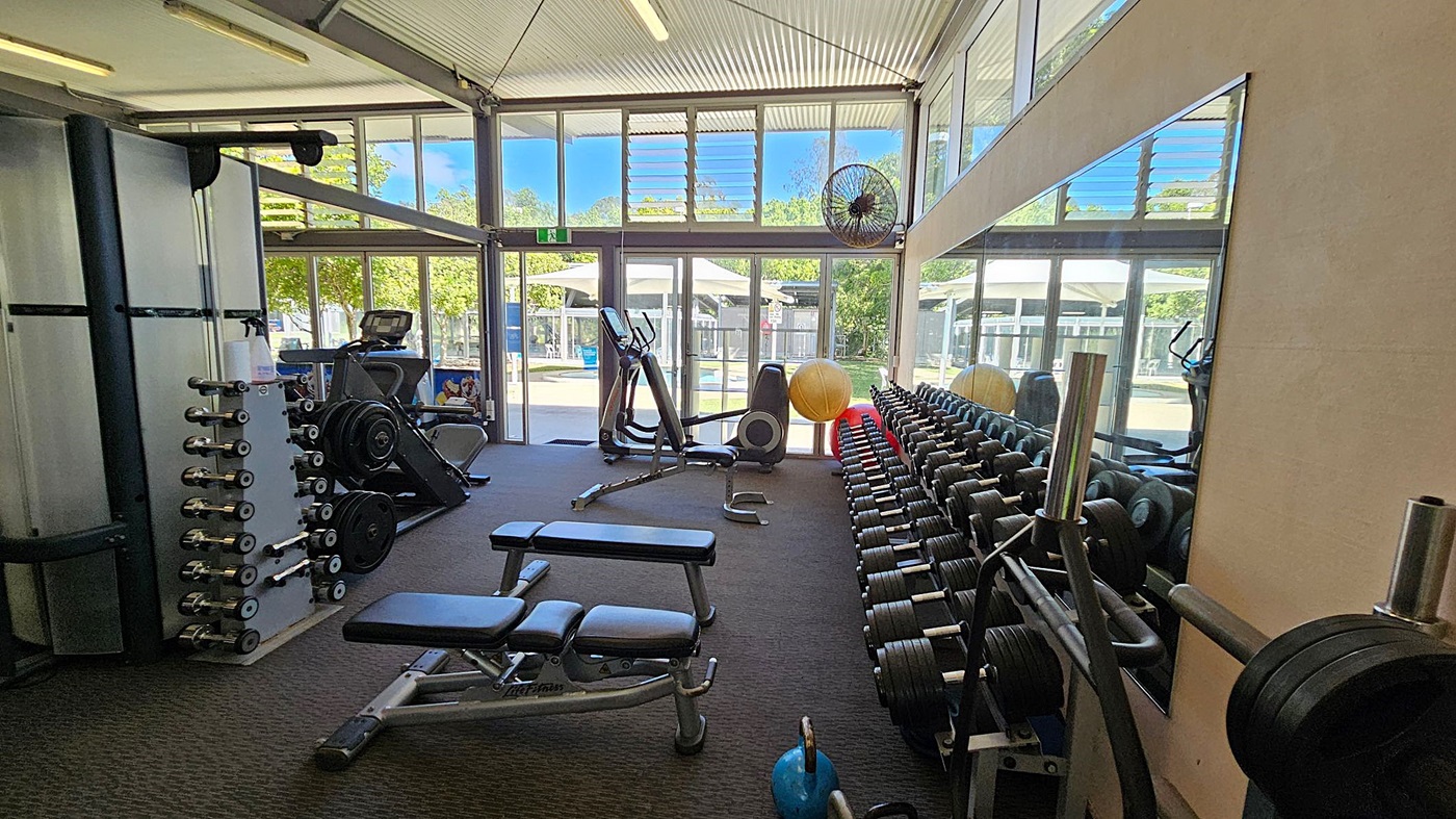 A gym with large windows and glass doors allowing natural light to enter. On the left, there is a weight machine and a rack of dumbbells. In the middle, there are two adjustable benches and a stationary bike. On the right, there is a long mirror reflecting the equipment and a row of dumbbells. A large yellow exercise ball and a blue kettlebell are also visible. The floor is carpeted, and the ceiling has exposed beams with fluorescent lights. A gym with large windows and glass doors allowing natural light to enter. On the left, there is a weight machine and a rack of dumbbells. In the middle, there are two adjustable benches and a stationary bike. On the right, there is a long mirror reflecting the equipment and a row of dumbbells. A large yellow exercise ball and a blue kettlebell are also visible. The floor is carpeted, and the ceiling has exposed beams with fluorescent lights.