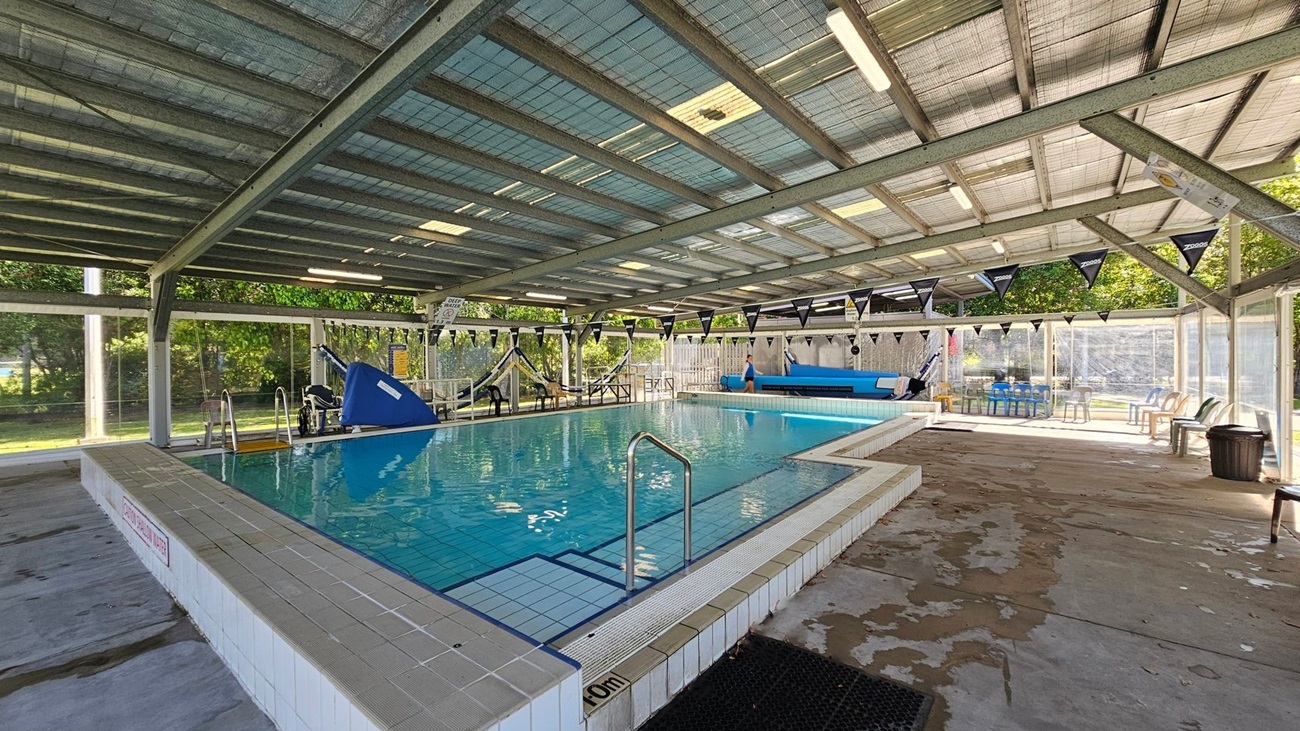 Indoor swimming pool with a tiled edge and clear water. The ceiling is made of metal beams with translucent panels, allowing natural light to filter through. On the left, there are large windows with a view of green trees and grass outside. On the right, there is pool equipment, including a blue pool cover roller. In the middle, a blue slide is visible at the far end of the pool. The floor around the pool is concrete with some wet patches. Black triangular flags are strung across the pool. Indoor swimming pool with a tiled edge and clear water. The ceiling is made of metal beams with translucent panels, allowing natural light to filter through. On the left, there are large windows with a view of green trees and grass outside. On the right, there is pool equipment, including a blue pool cover roller. In the middle, a blue slide is visible at the far end of the pool. The floor around the pool is concrete with some wet patches. Black triangular flags are strung across the pool.